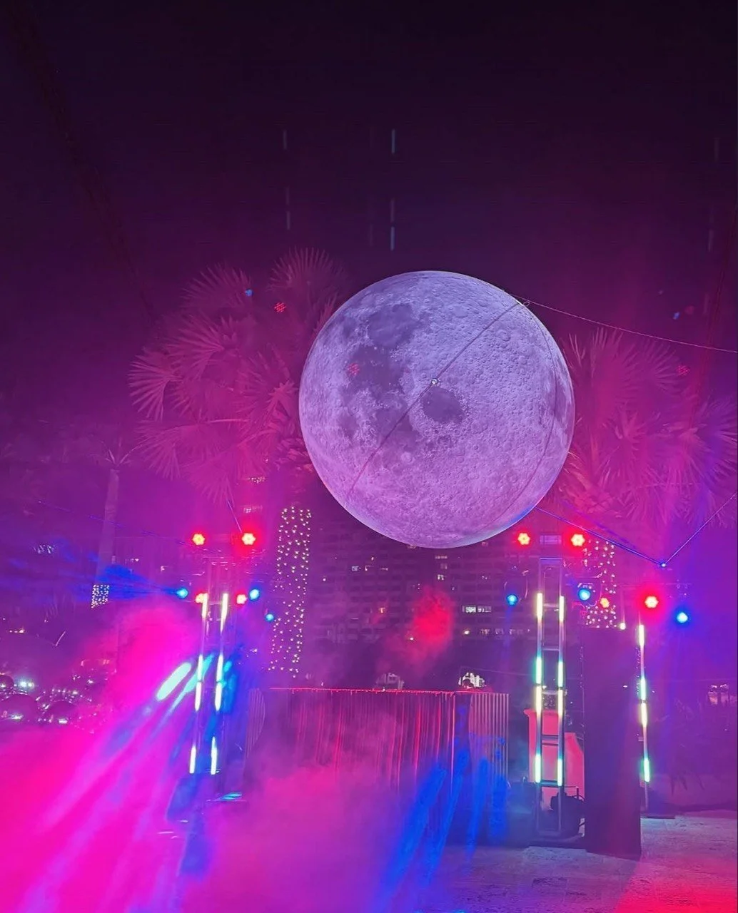 A large illuminated moon decoration hanging in the night sky with colorful stage lights and palm trees in the background, creating a vibrant party atmosphere.