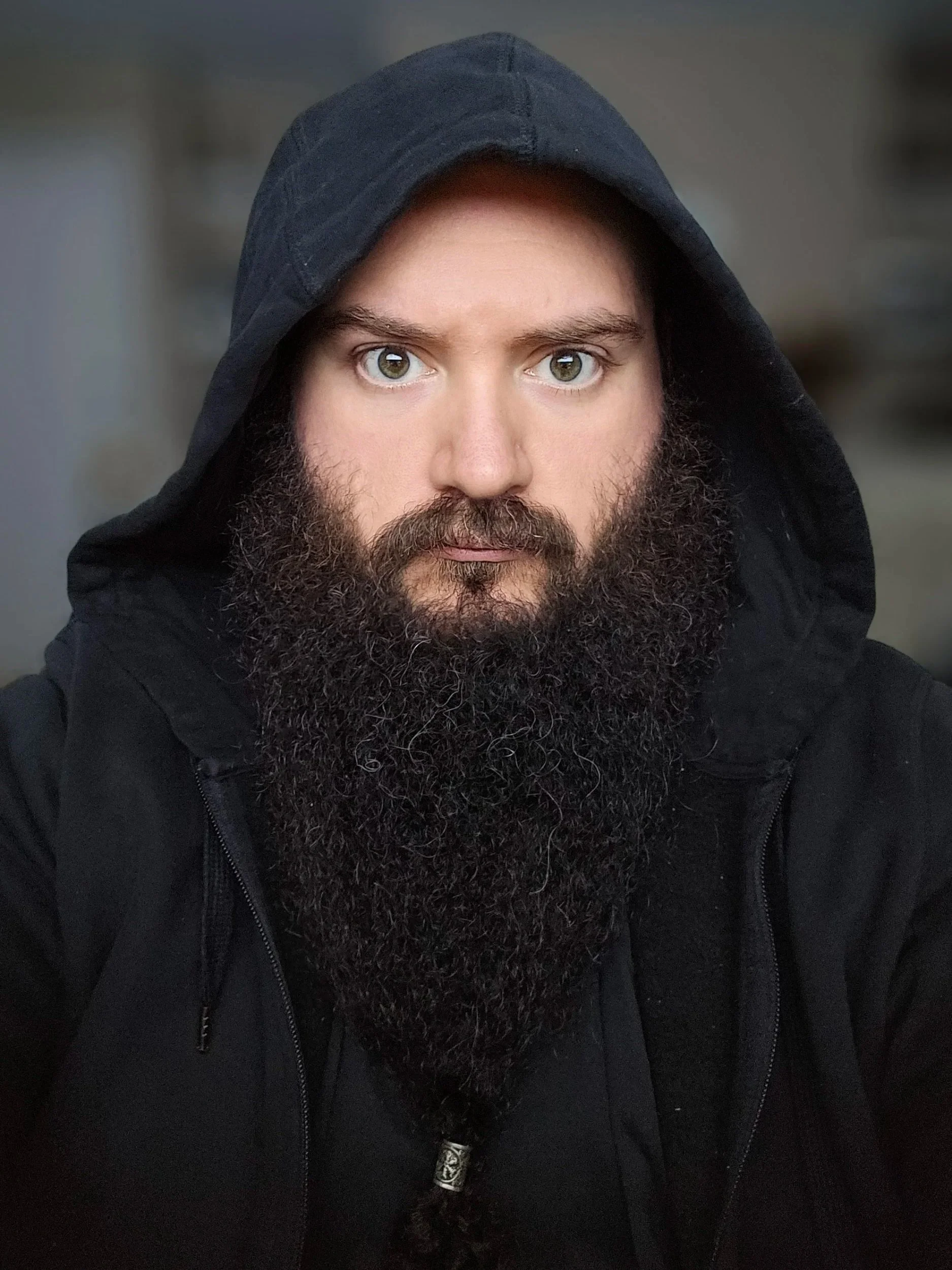 A man with a thick beard and mustache wearing a dark hoodie, looking directly at the camera.