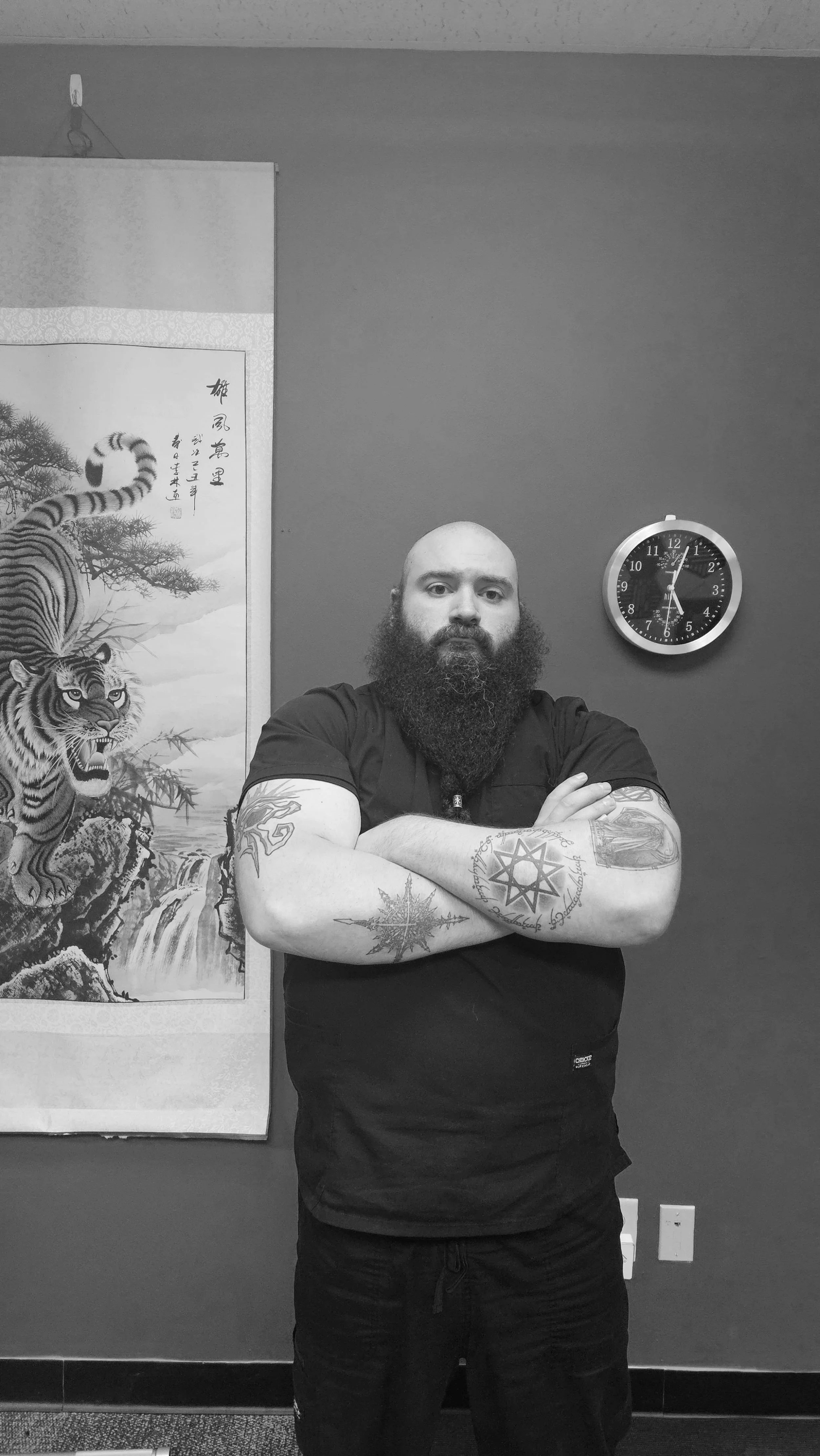 A man with a beard and tattoos on his arms standing with arms crossed in front of a wall with traditional Asian artwork and a clock showing 1:00.
