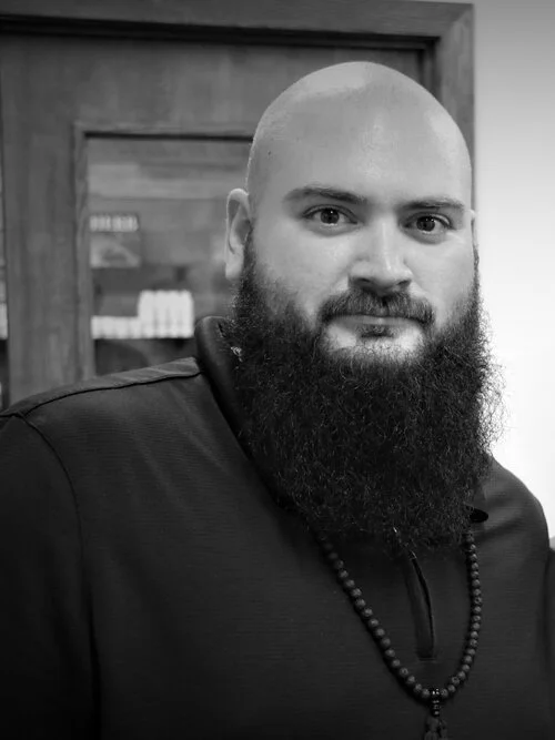 A man with a shaved head, beard, and mustache wearing a black shirt and a beaded necklace, standing in front of a wooden cabinet with books or products inside.