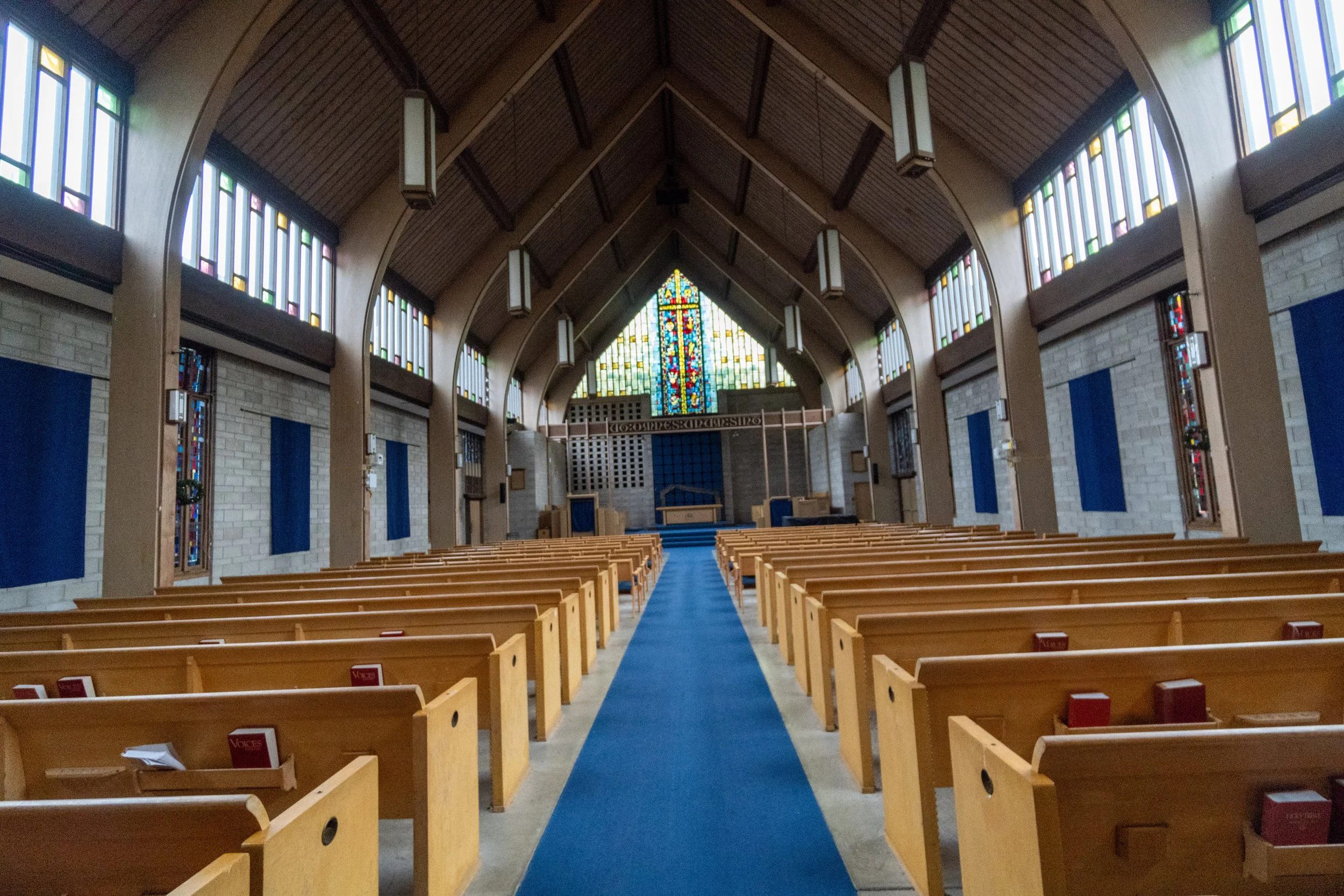 Interior of a church with wooden pews, a blue carpet down the aisle, a large stained glass window at the front, and hanging light fixtures, with side windows featuring colorful stained glass.