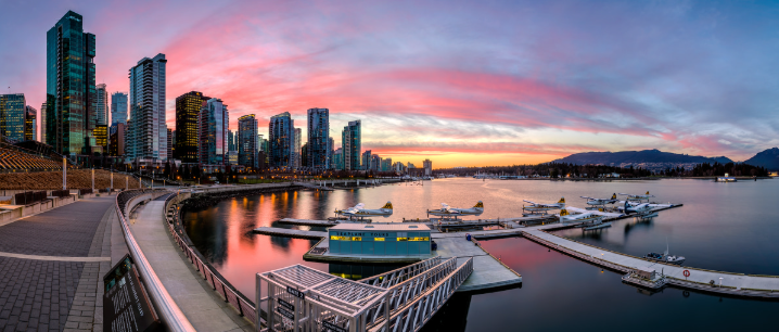 City skyline with tall modern buildings at sunset, waterfront with boats docked at a marina, and a paved walkway along the water.