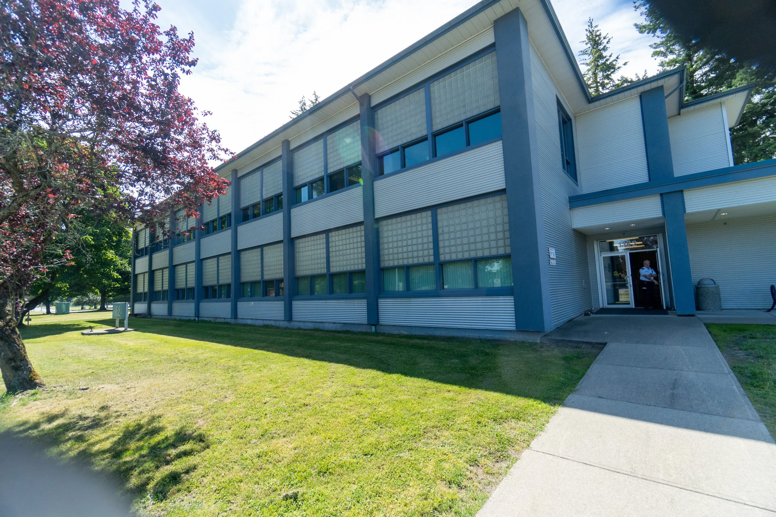 Exterior of a modern apartment building with a sidewalk leading to the entrance, surrounded by green grass and trees, one with red leaves, on a clear day.