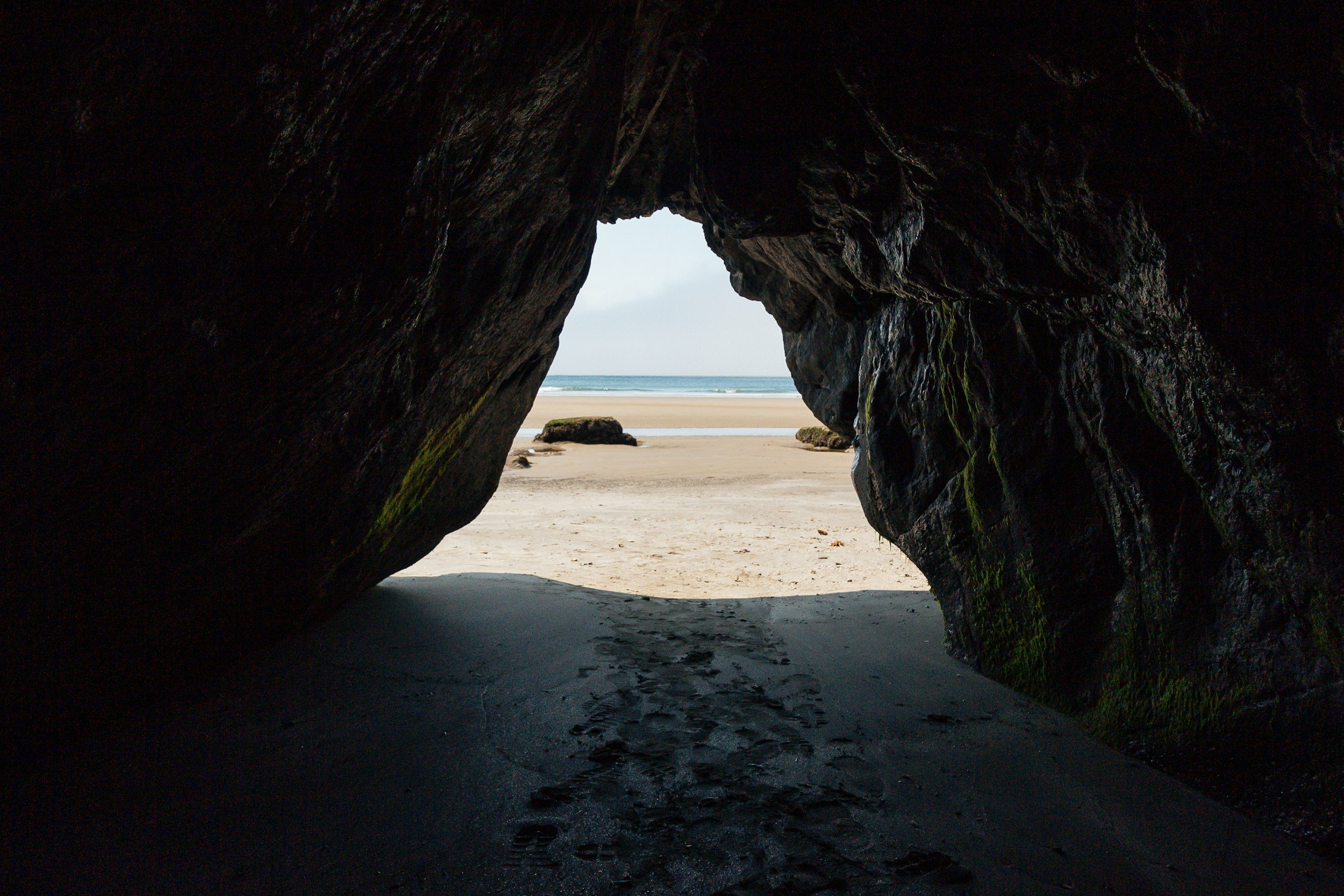 View of a sandy beach through a rocky cave opening with footprints in the sand, rocks in the distance, and the ocean beyond.