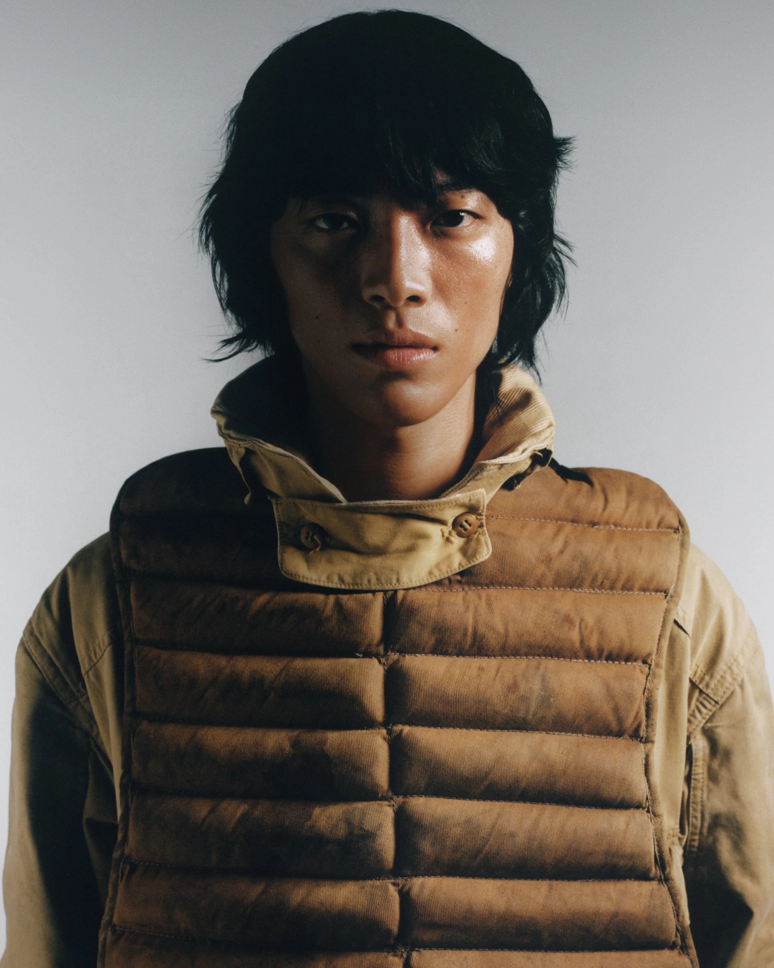 Un homme au regard sérieux portant une veste marron avec un col relevé, fond neutre.