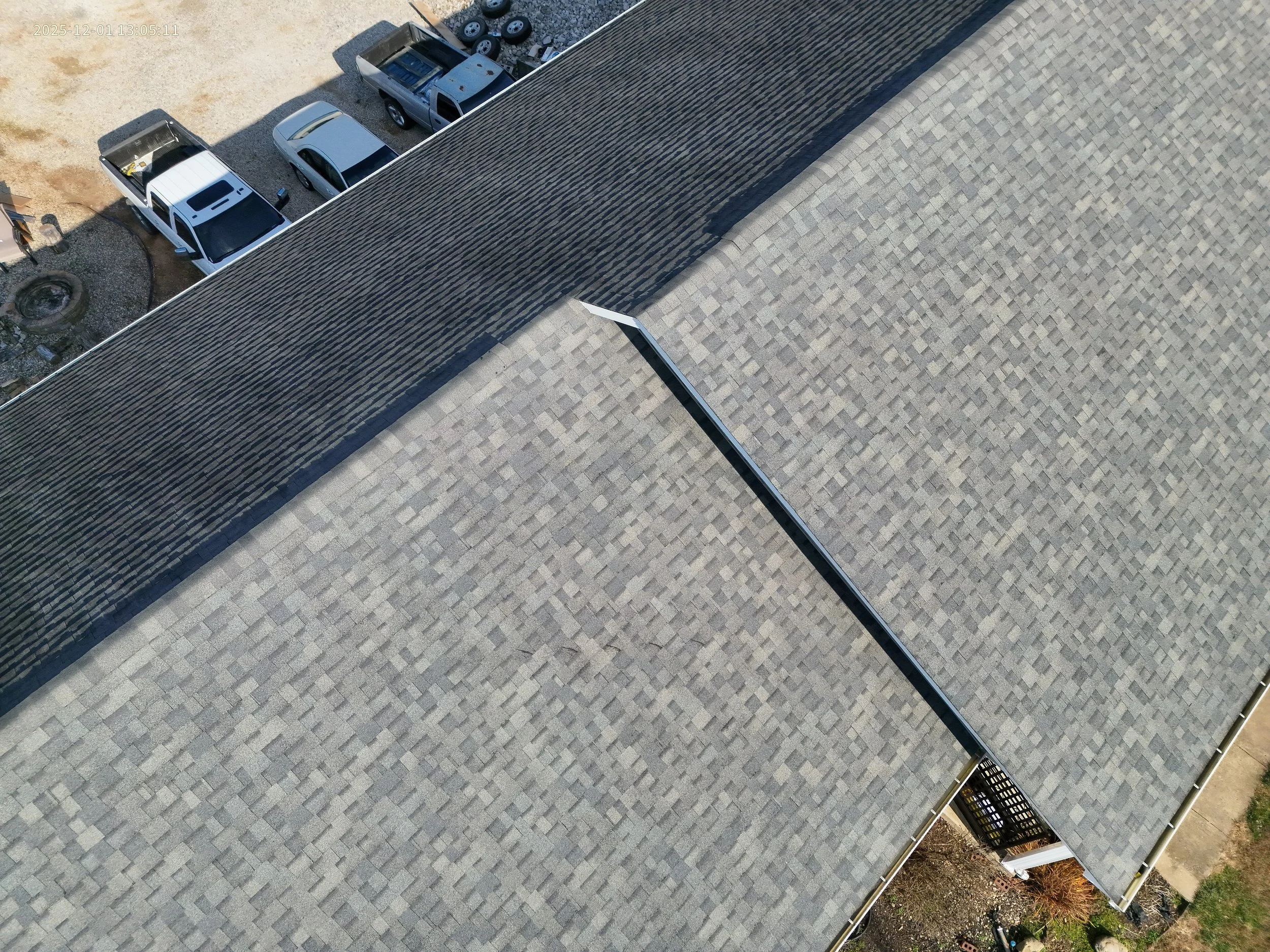 An aerial view of a building's roof with two different sections, one with darker shingles and one with lighter shingles, and a parking lot with several parked cars nearby.