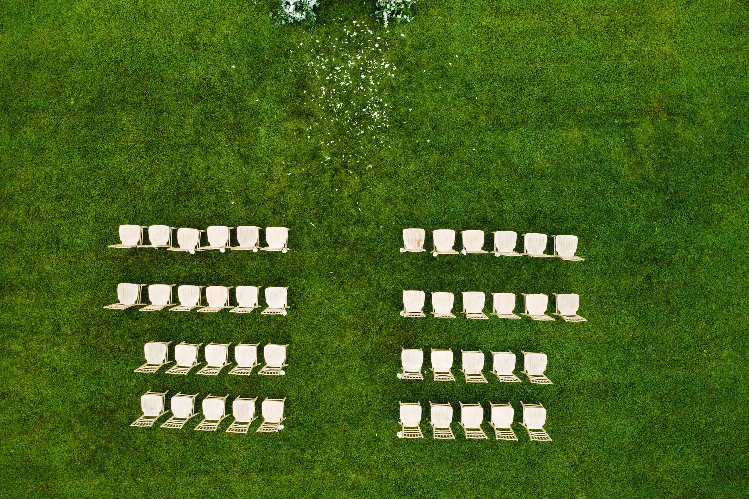 An aerial view of many white folding chairs arranged on a grassy field, with some scattered flower petals and two rows of chairs facing each other. Likely a wedding ceremony.