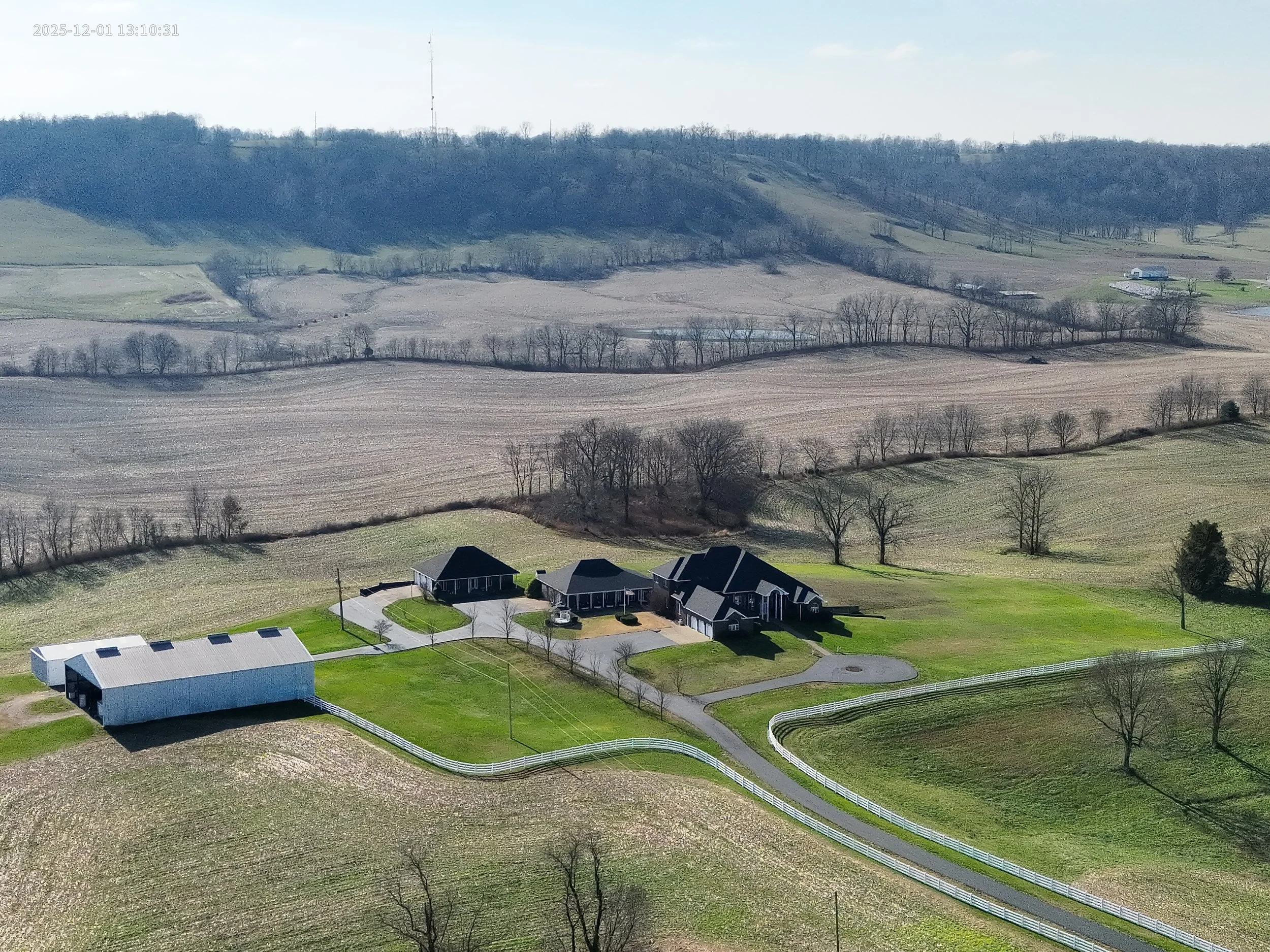 A rural farmstead with a large house, several outbuildings, a circular driveway, and a white fence in a green field, surrounded by rolling hills and farmland.