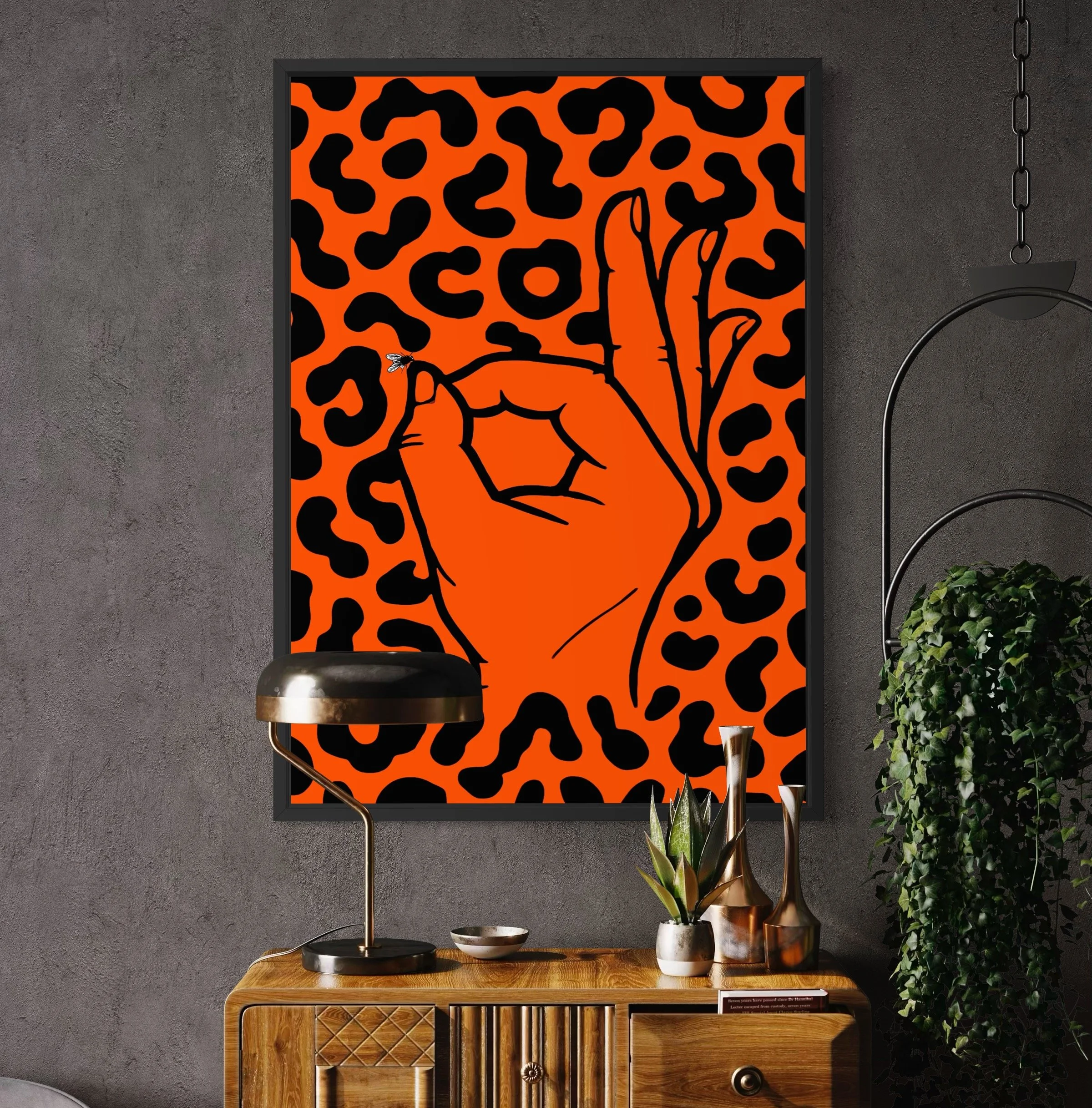 A framed abstract art print of an orange hand making the 'OK' gesture against a bright orange and black leopard print background hangs on a textured gray wall. A wooden sideboard with decorative items. Street art from London