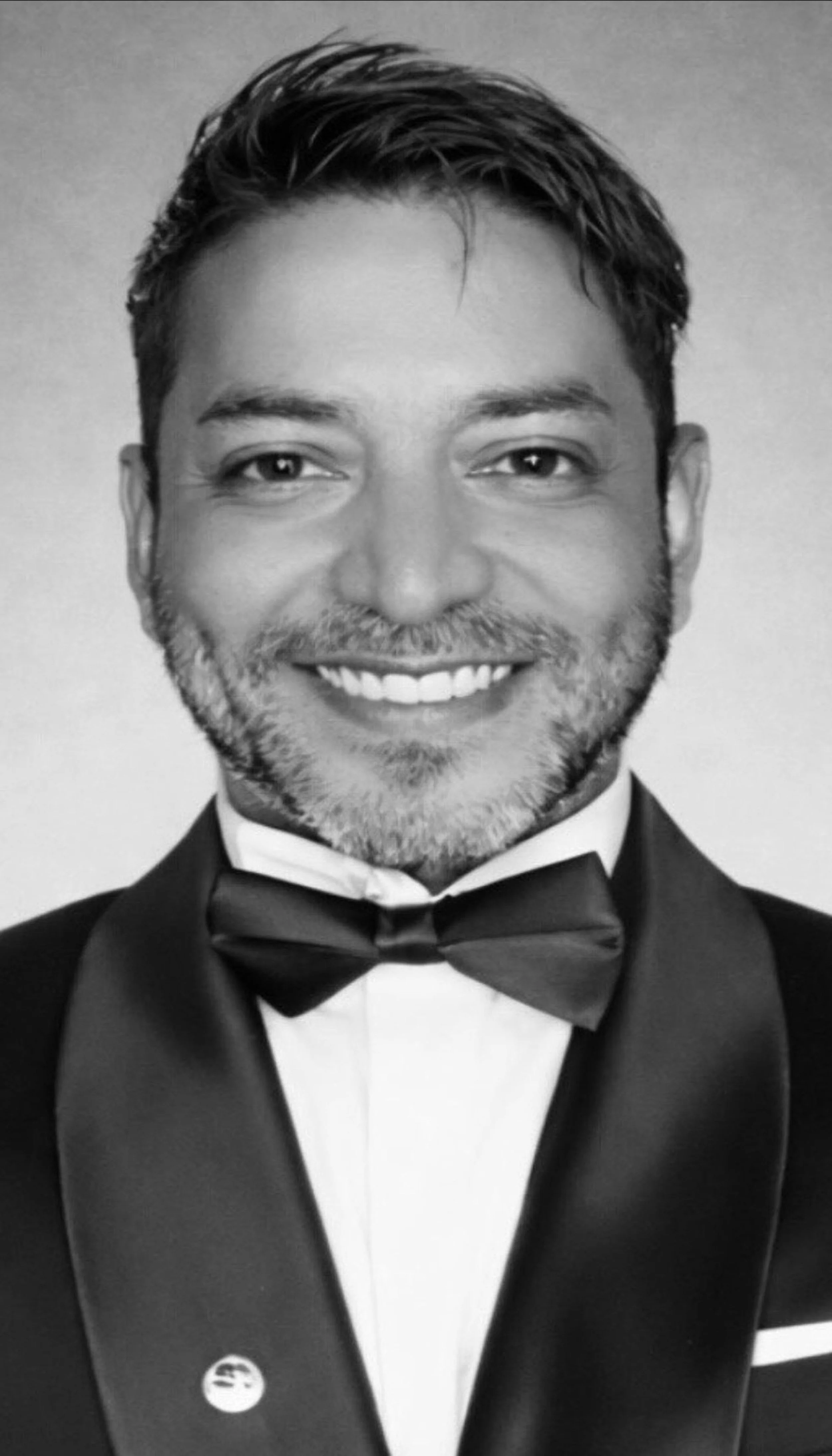 Black and white portrait of a man with short, dark hair, smiling, dressed in a tuxedo with a bow tie, against a plain background.