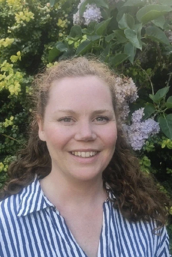 A woman with curly brown hair smiling outdoors, surrounded by green foliage and purple and light pink hydrangea flowers.