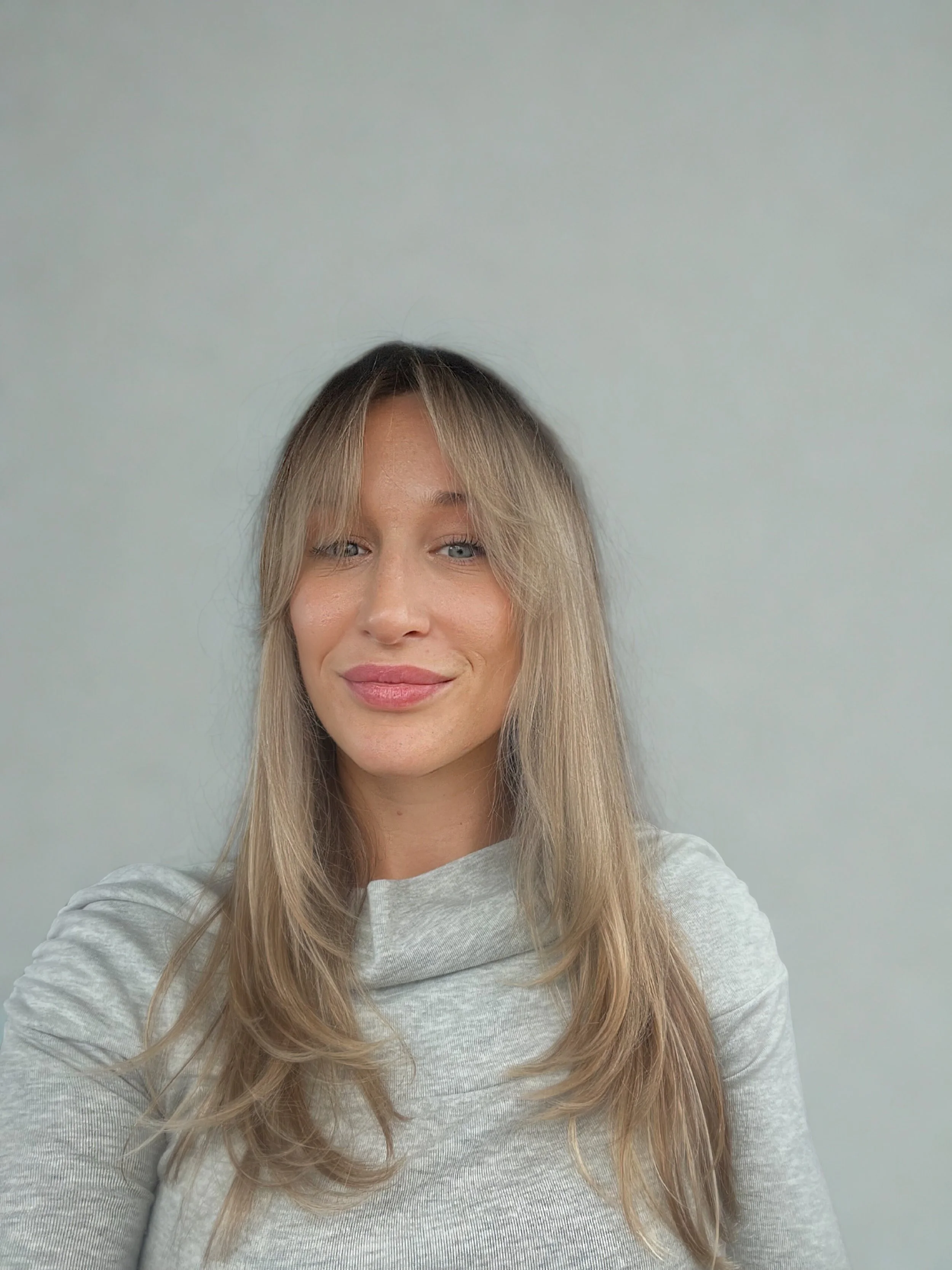 A woman with blonde hair and blue eyes taking a selfie against a plain light-colored background, wearing a light gray top.