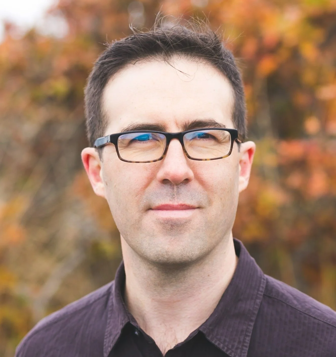 A man with glasses standing outdoors with autumn-colored trees in the background.