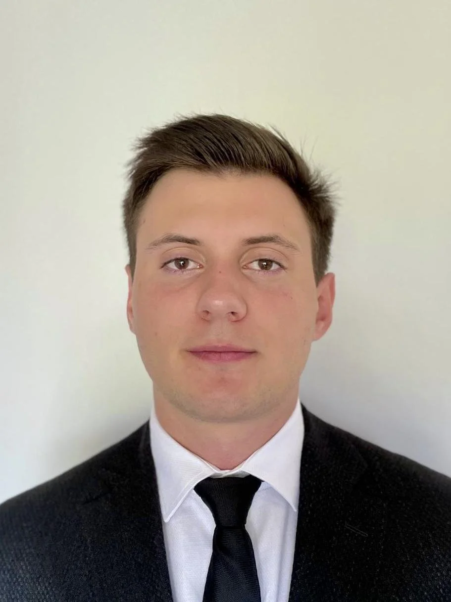 Portrait of a young man with short brown hair, wearing a black suit, white shirt, and black tie, standing against a plain off-white background.