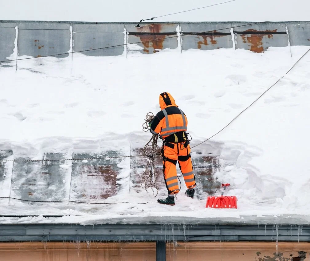 Un travailleur en équipement de sécurité orange et noir en train de couper ou de travailler sur une pièce de métal recouverte de neige et de glace sur un toit.