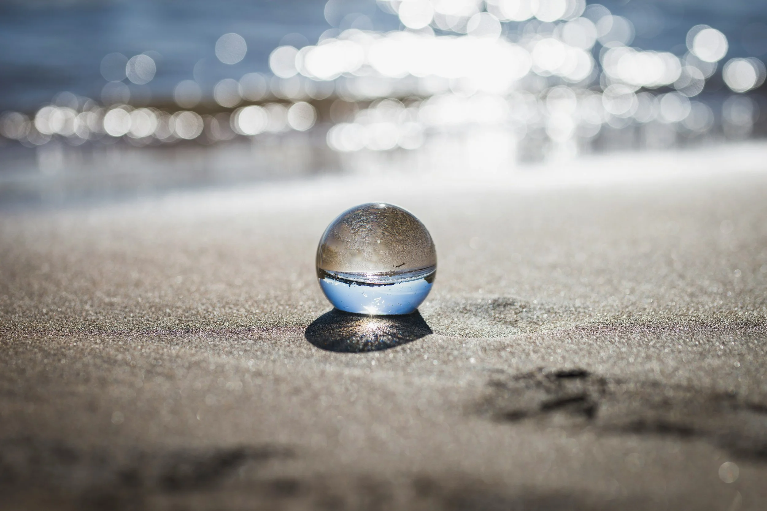 A clear glass sphere resting on the sand at a beach with sunlight reflecting off the water in the background, creating a bokeh effect.