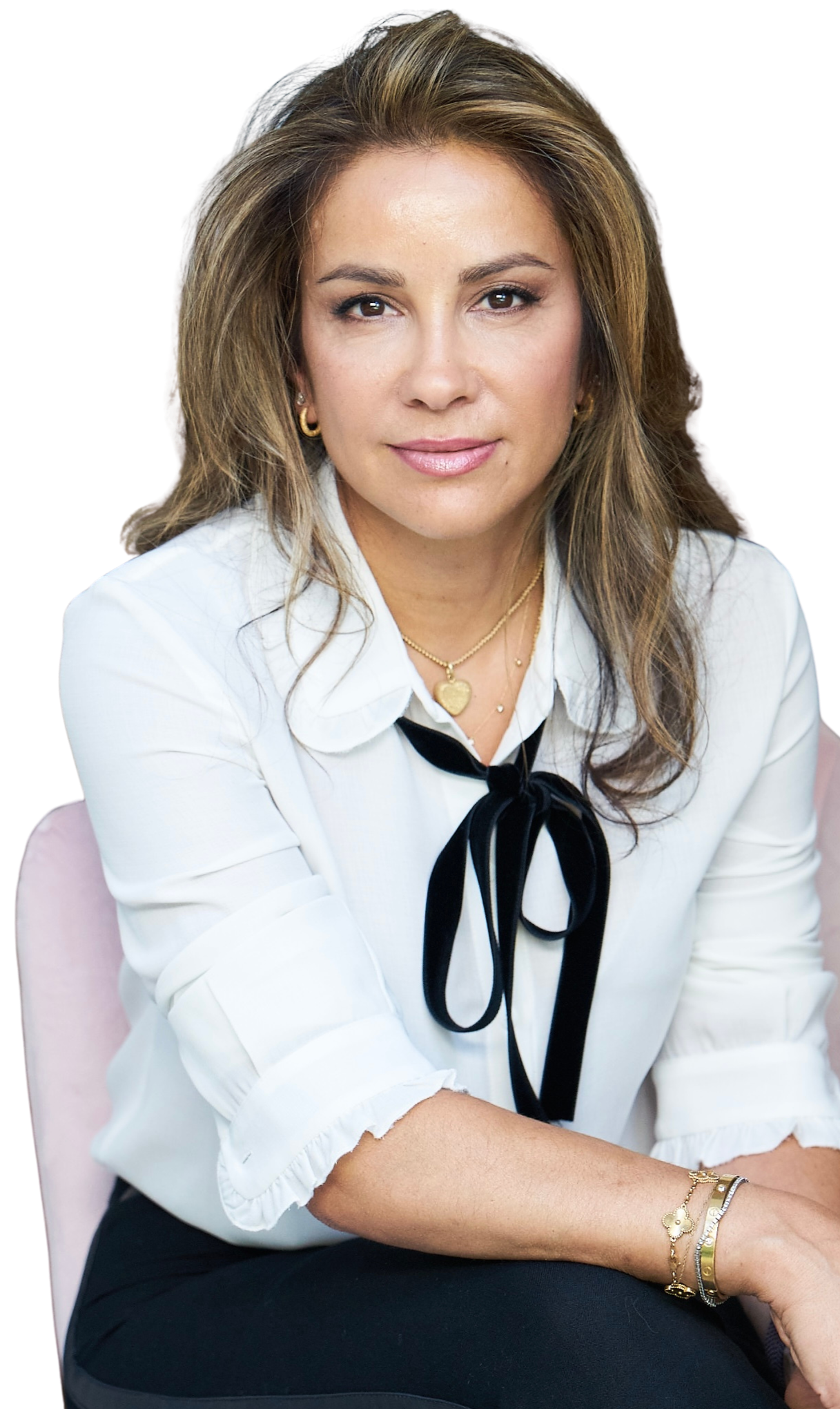 A woman with light brown hair and tan skin sits with her arms crossed. She wears a white blouse with a black bow at the collar, gold earrings, a gold necklace with a heart pendant, and gold and silver bracelets. She is smiling softly and looking directly at the camera.