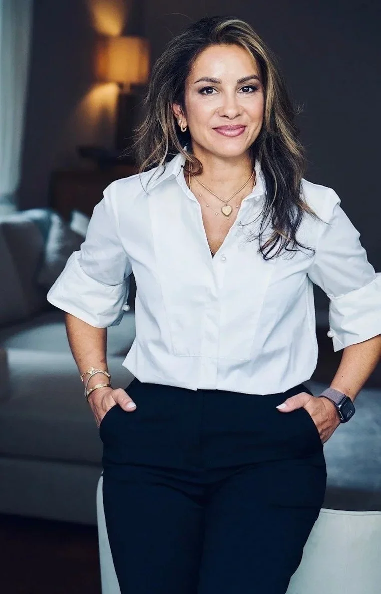 A woman with long, wavy hair wearing a white blouse and black pants, standing confidently in a cozy, dimly lit living room with a sofa and a lamp in the background.