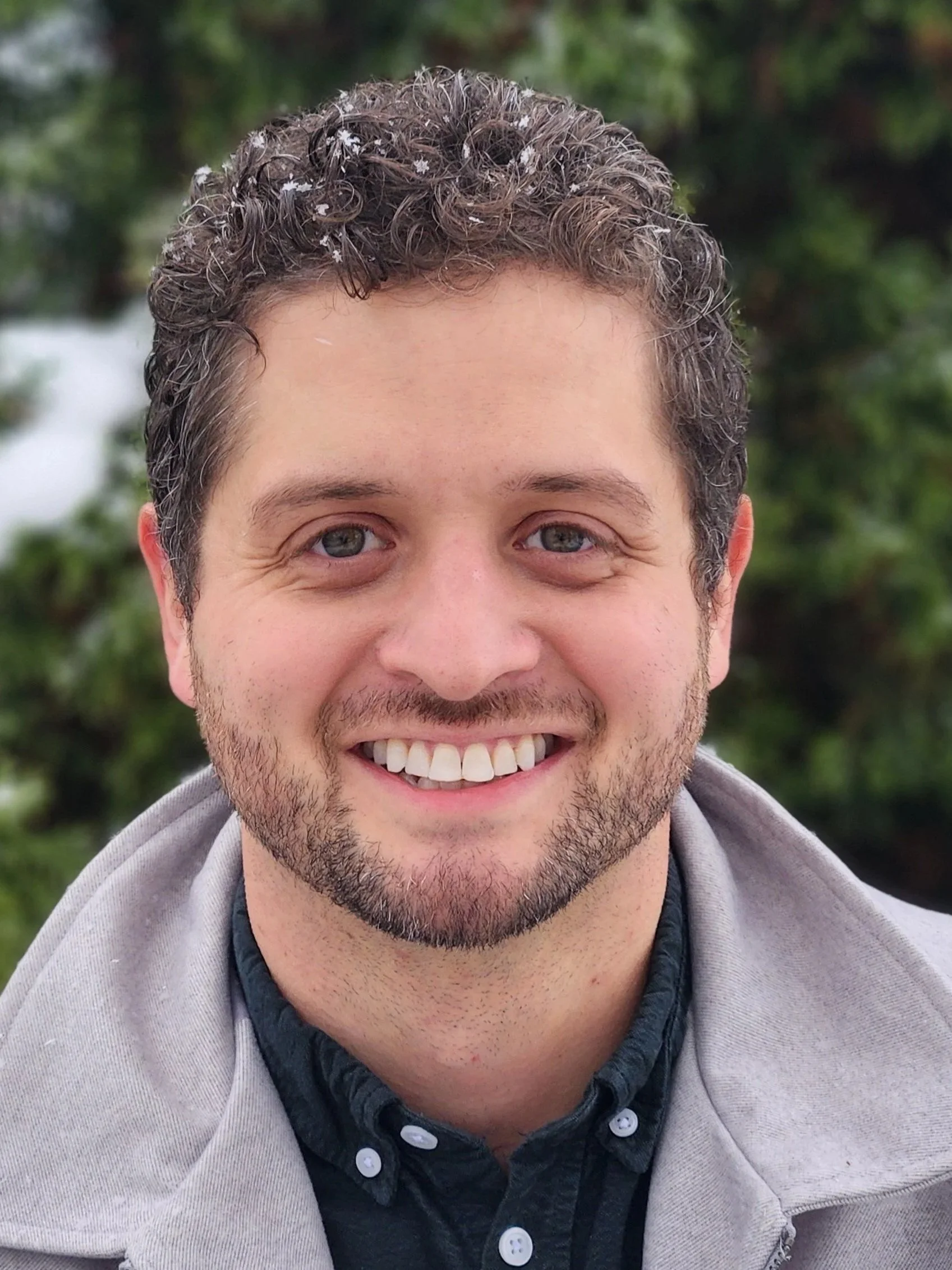 Close-up of smiling man with short curly hair and blue eyes, outdoors with blurred green foliage background.