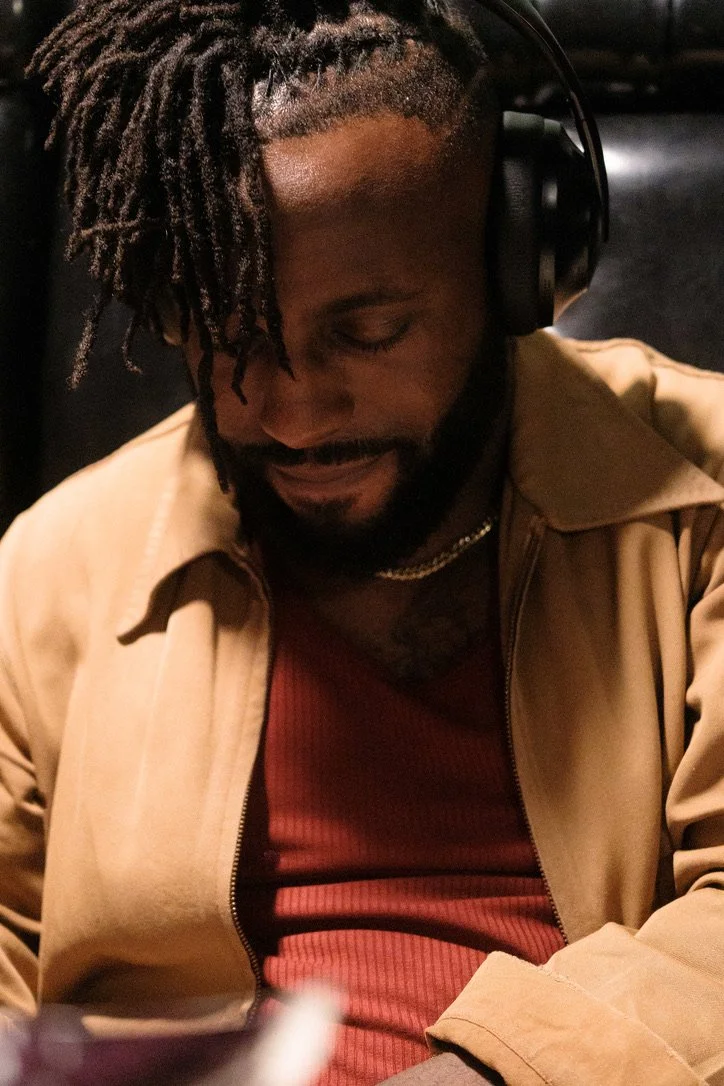 Brunson is wearing headphones, a beige jacket, and a red shirt, smiling with eyes closed.