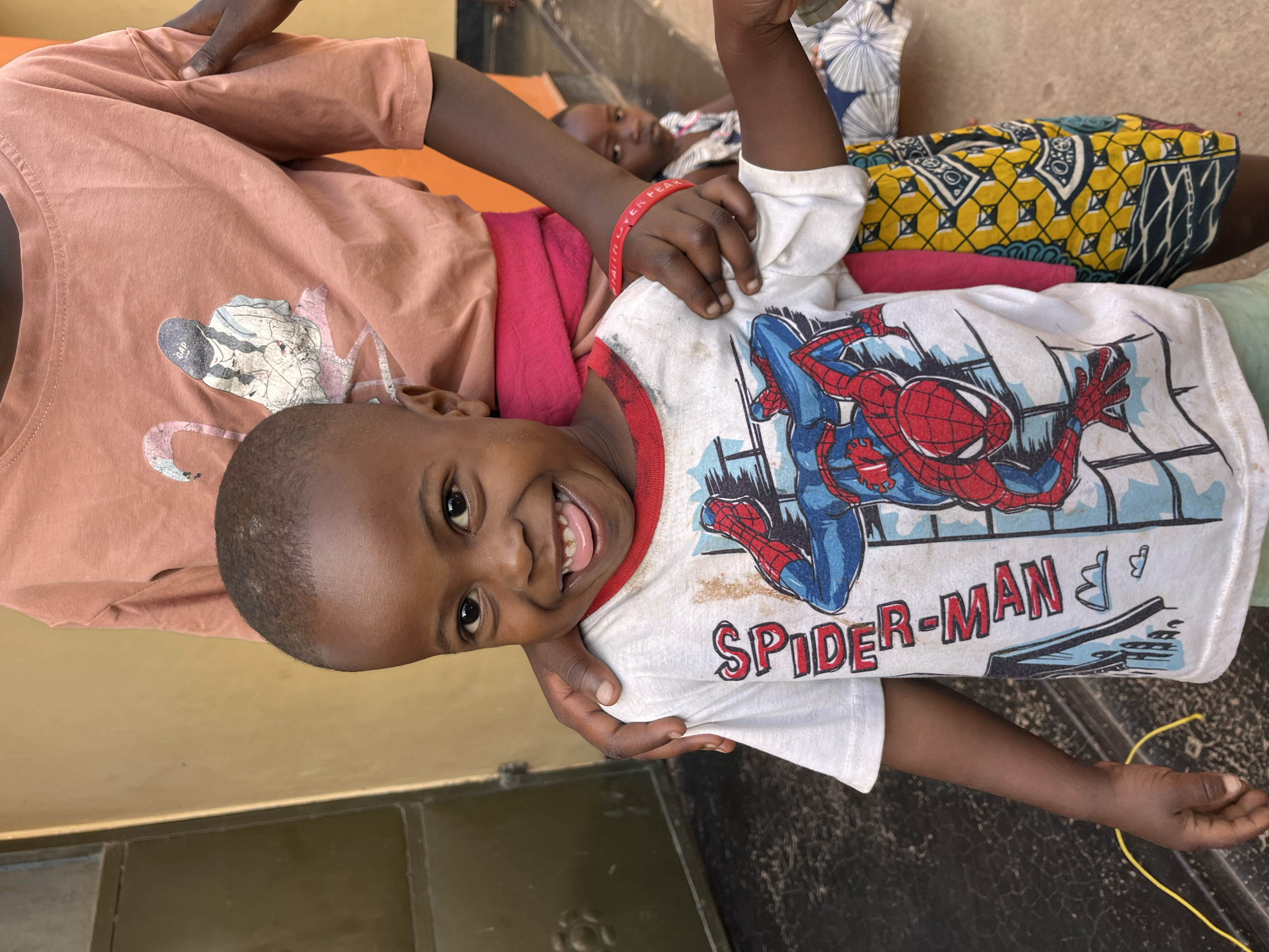 A young boy smiling and holding up a peace sign with his fingers, wearing a Spider-Man t-shirt, and sitting next to a woman. In the background, another person is lying on a bed or couch.