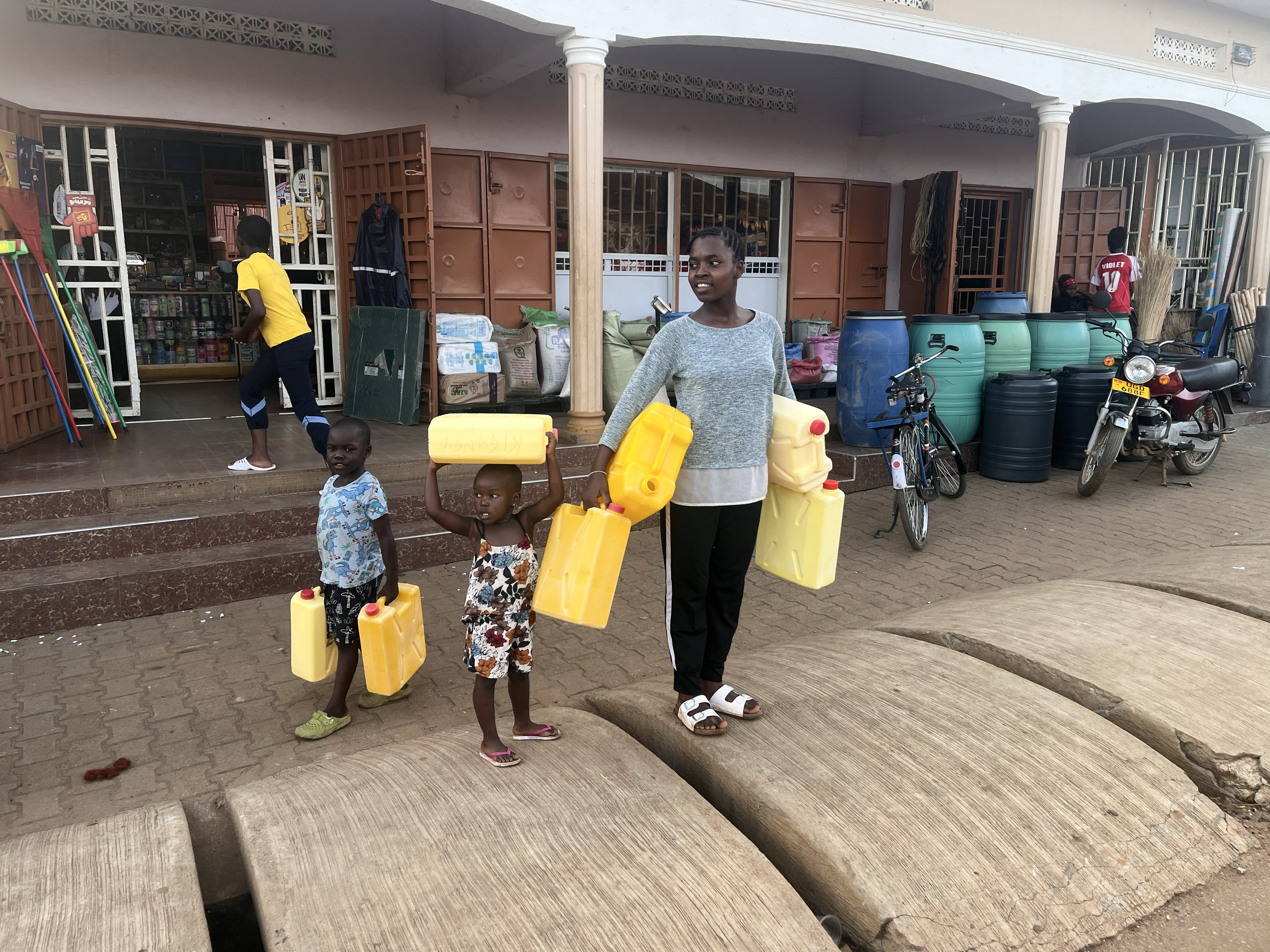 A woman and two children carrying yellow water containers walking outside a shop. The shop has open doors displaying shelves of goods, and bicycles and motorcycles parked outside.