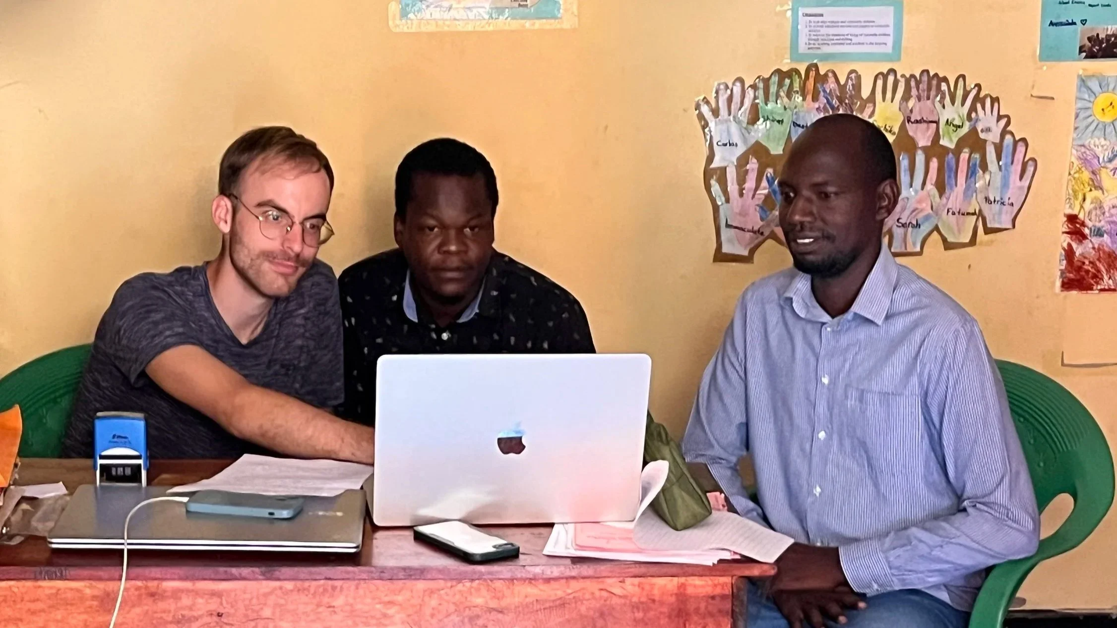Three men sitting at a desk with a laptop and papers, working together in an office or classroom setting, with colorful artwork and hand cutouts on the wall behind them.