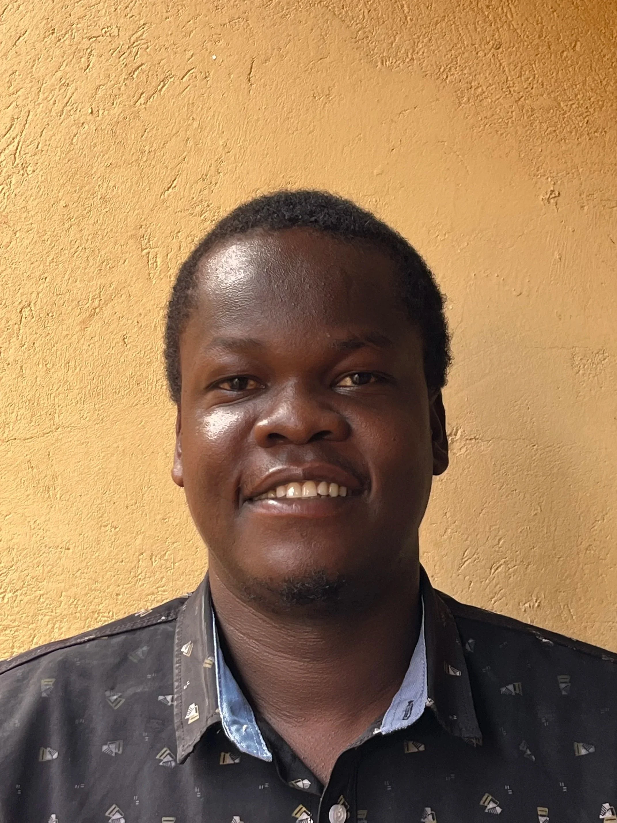 Portrait of a smiling young man with dark skin and short black hair, wearing a black shirt with patterned details, standing against a yellow textured wall.