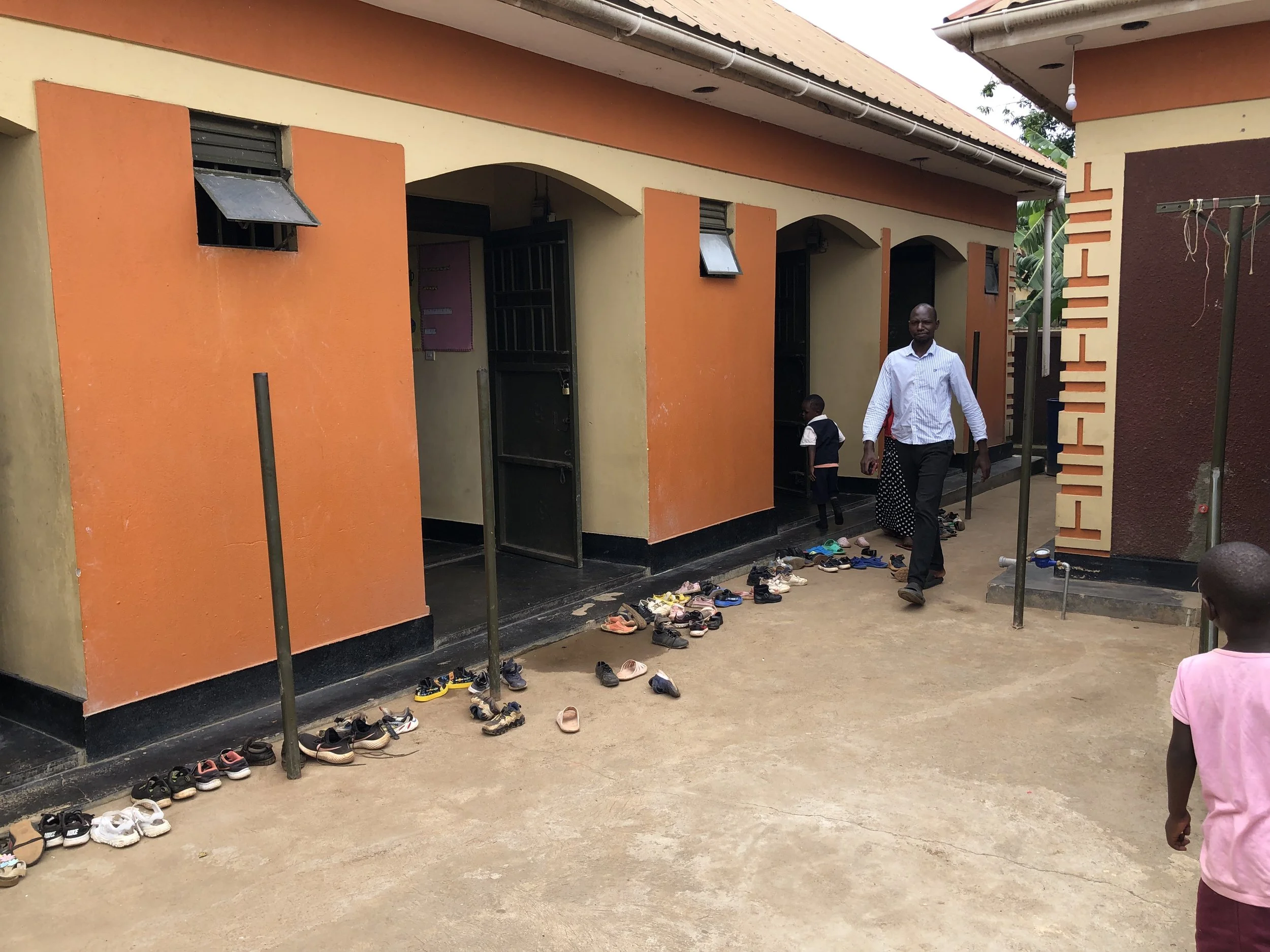 A school courtyard with shoes lined up outside classroom doors. A man and two children are present, one child wearing a school uniform. The school building has orange and beige walls with black doors, and the word 'CHILD' is visible on a sign.