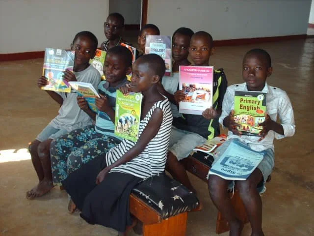 Seven children sitting on benches, each holding open books in a classroom or learning environment.