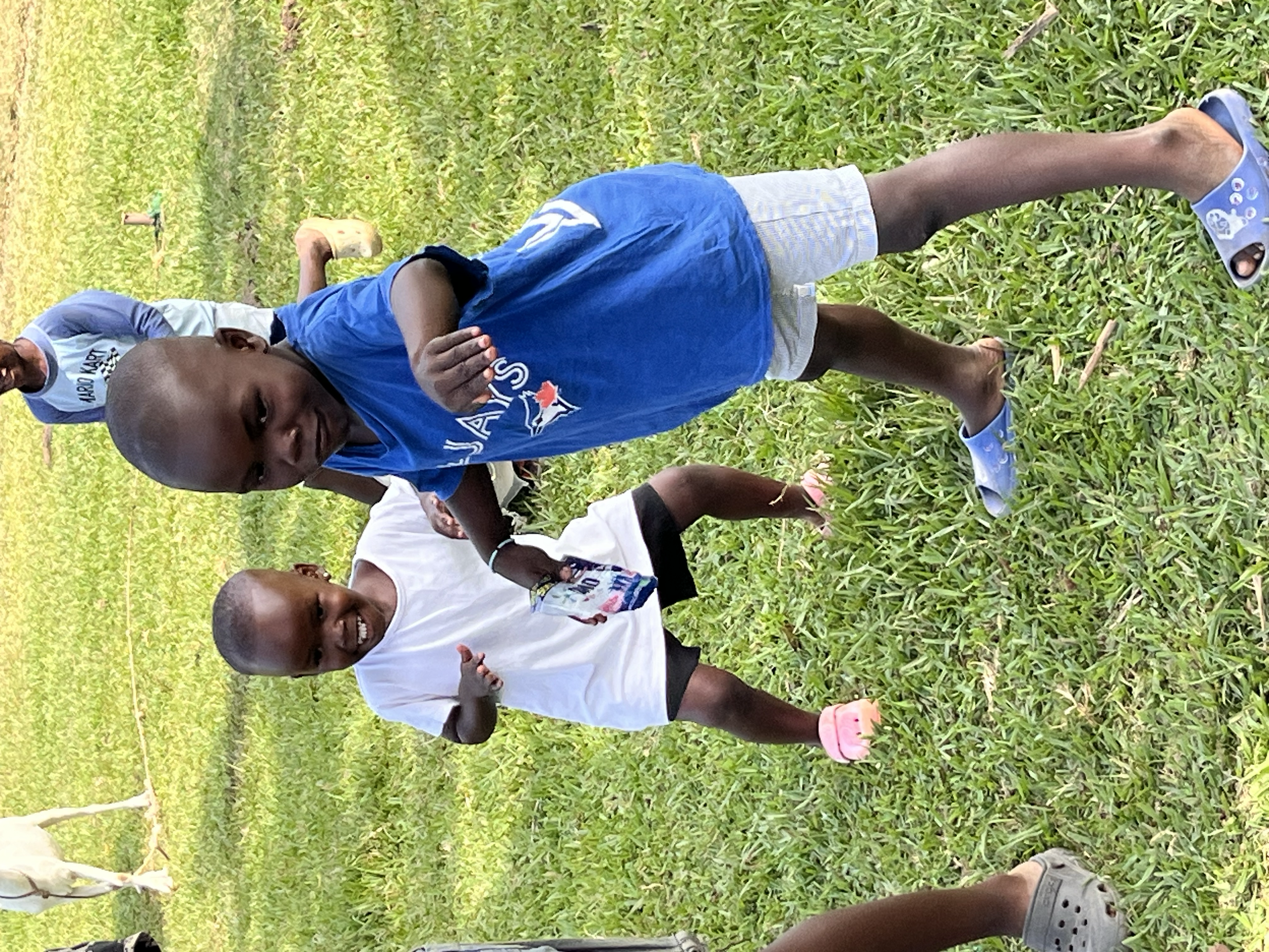 Two young boys with shaved heads playing on a grassy field. One is wearing a blue Toronto Blue Jays T-shirt, light-colored shorts, and blue sandals. The other boy is in a white T-shirt with black shorts and pink sandals. They are smiling and appear t