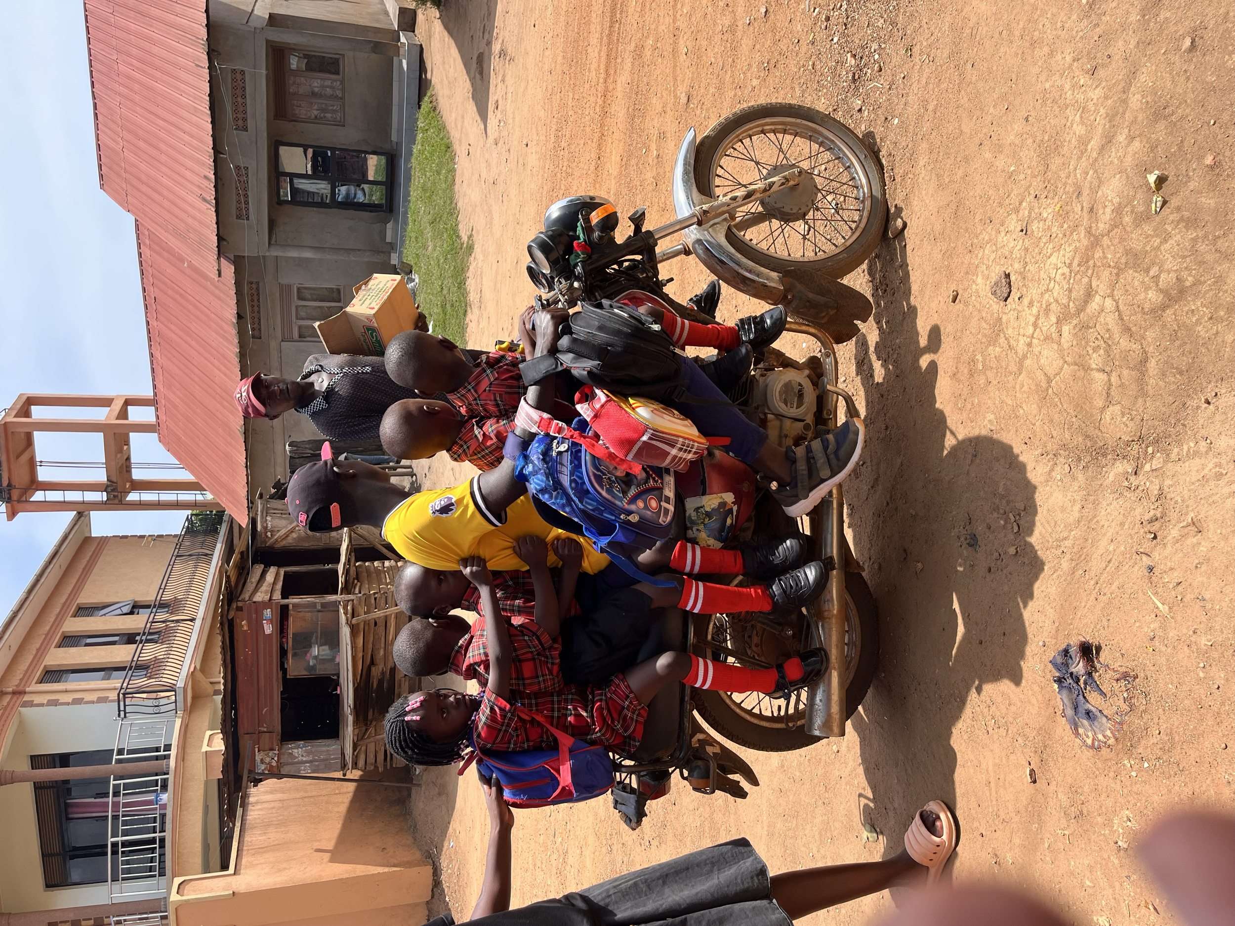 A group of children riding on a makeshift motorcycle tied to a tricycle in front of houses in a rural area.