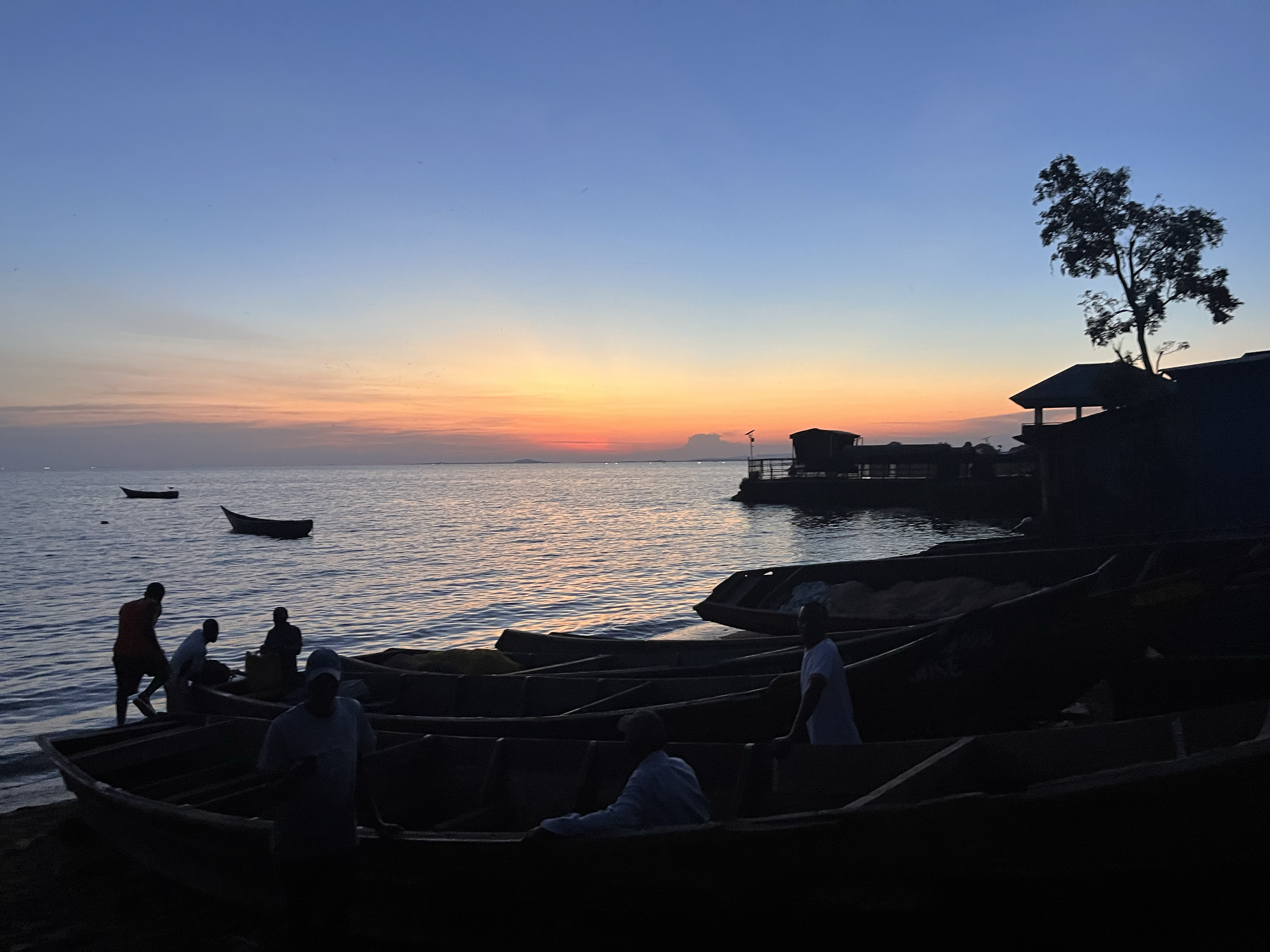 People at a beach during sunset with small boats on the water and a tree and building silhouette on the right.