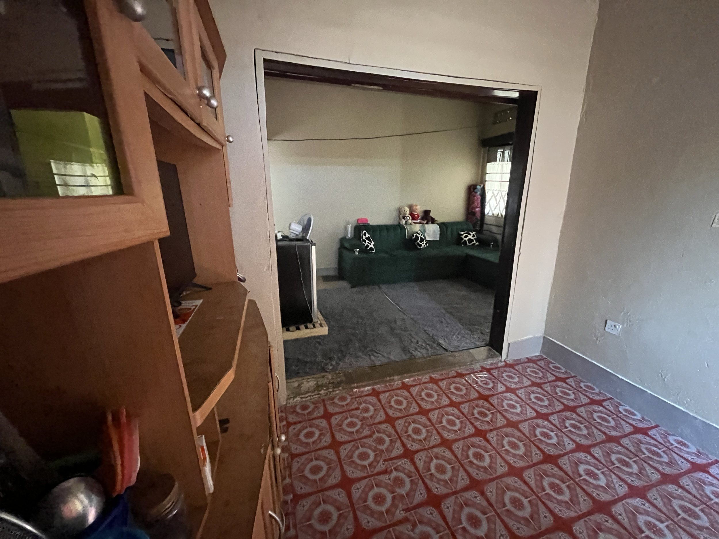 View of a living room through an open doorway with a patterned red and white floor in the foreground, a beige wall, a green sofa with black and white pillows, a small table with toys and a black portable heater, and partial window with curtains in th
