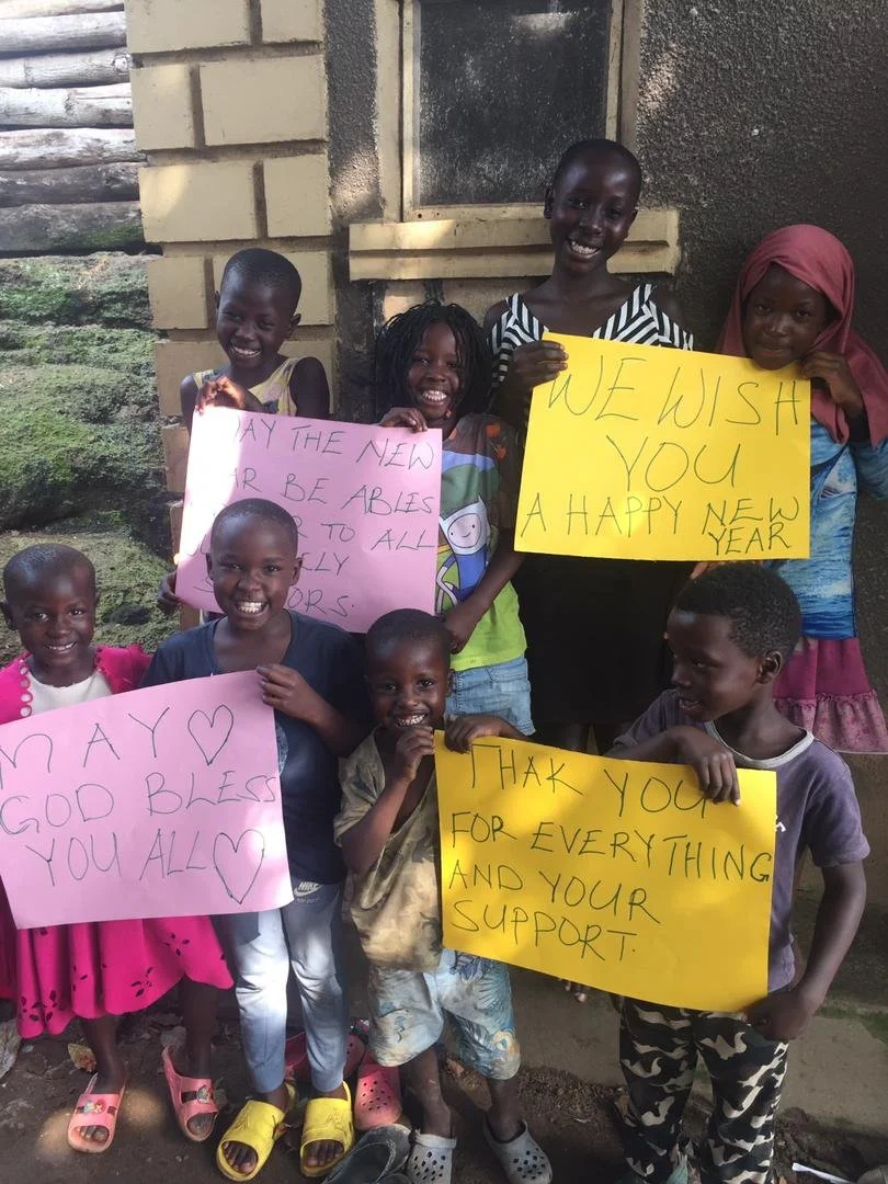Group of smiling children holding colorful signs with New Year wishes, standing outside near a building with brick wall and window.
