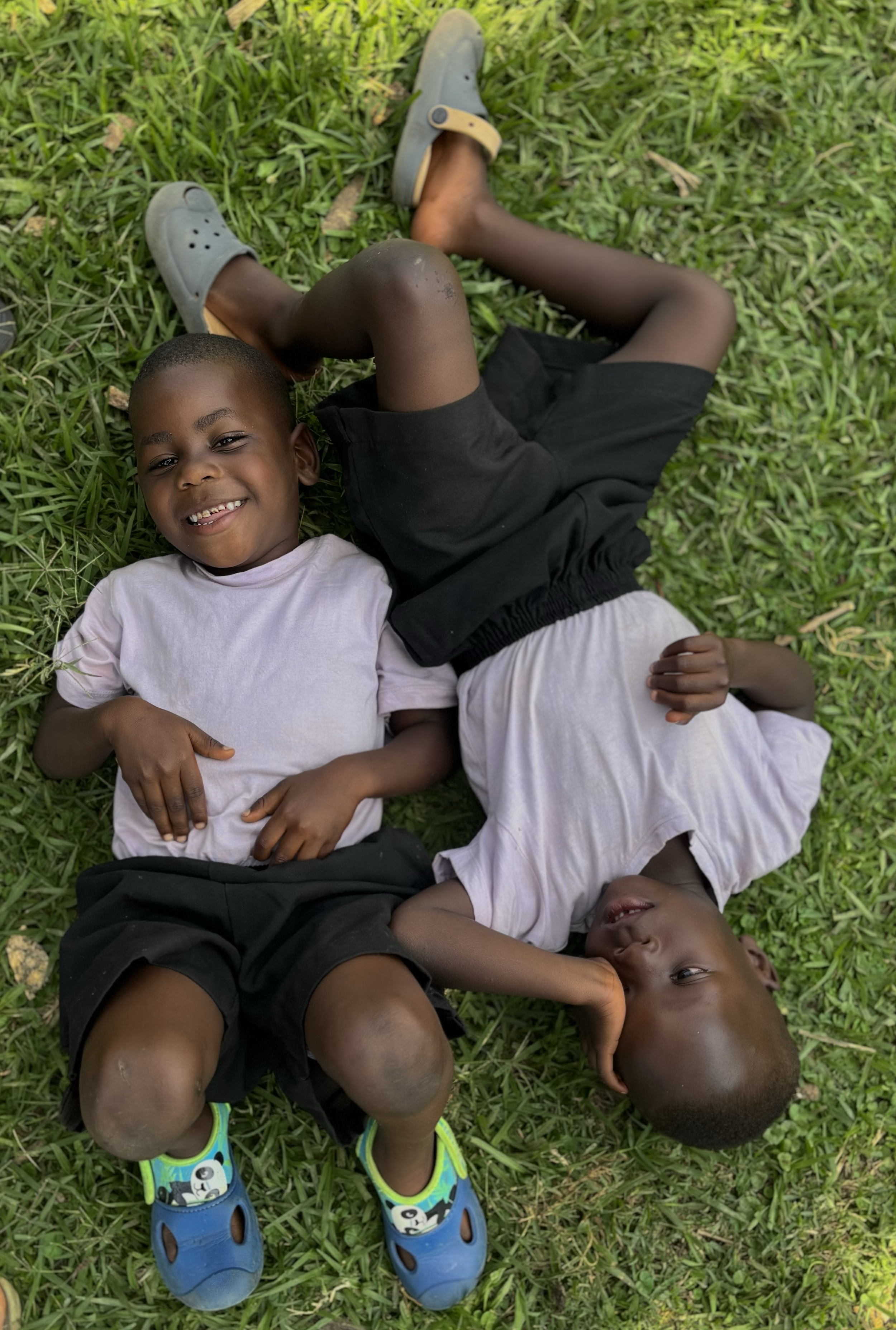 Two young boys lying on the grass, smiling, wearing casual clothes, with one boy wearing blue shoes with panda designs.