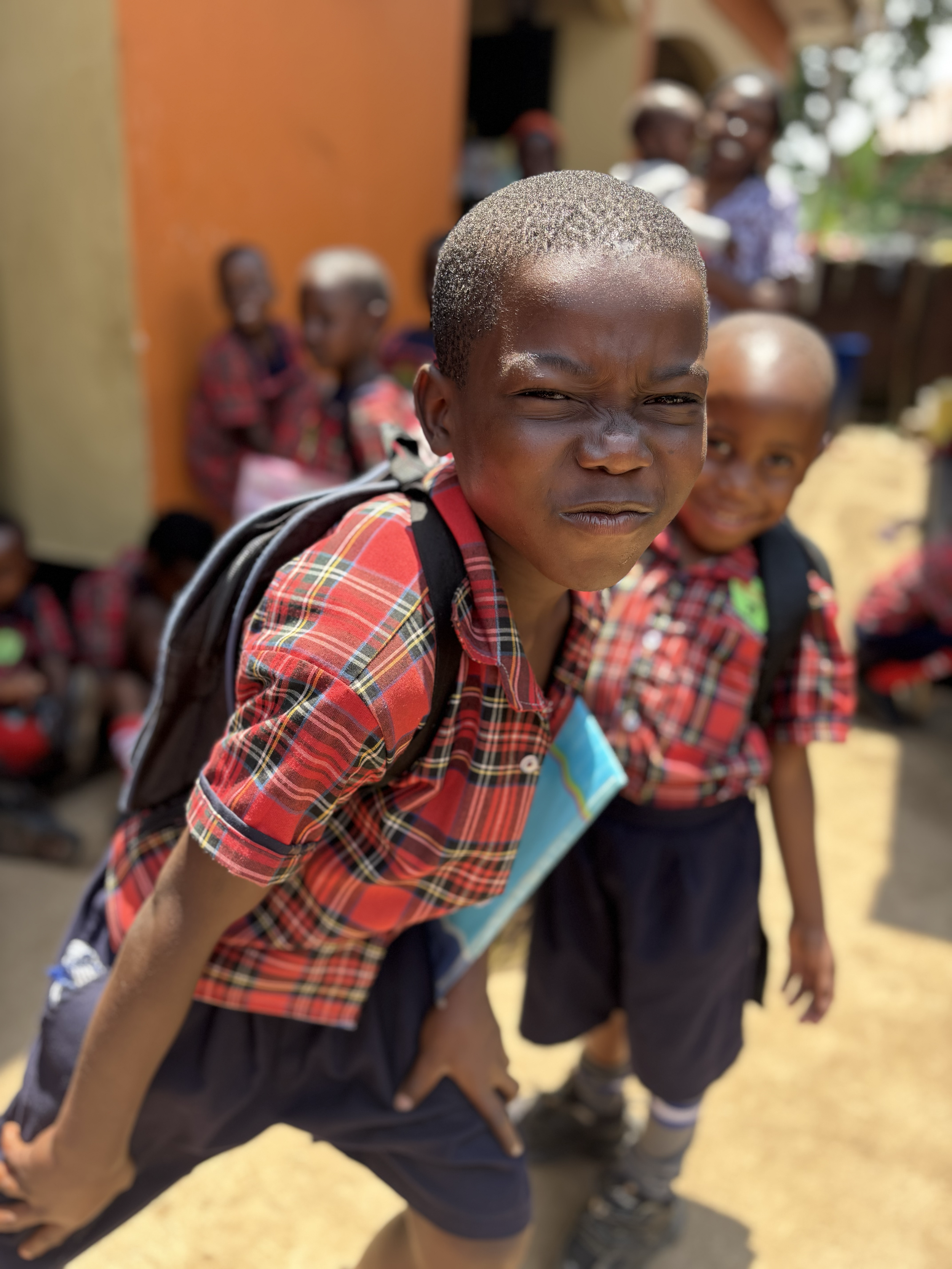 A young boy with a short haircut and a scrunched face, wearing a red plaid shirt and a backpack, standing outdoors with other children in the background.