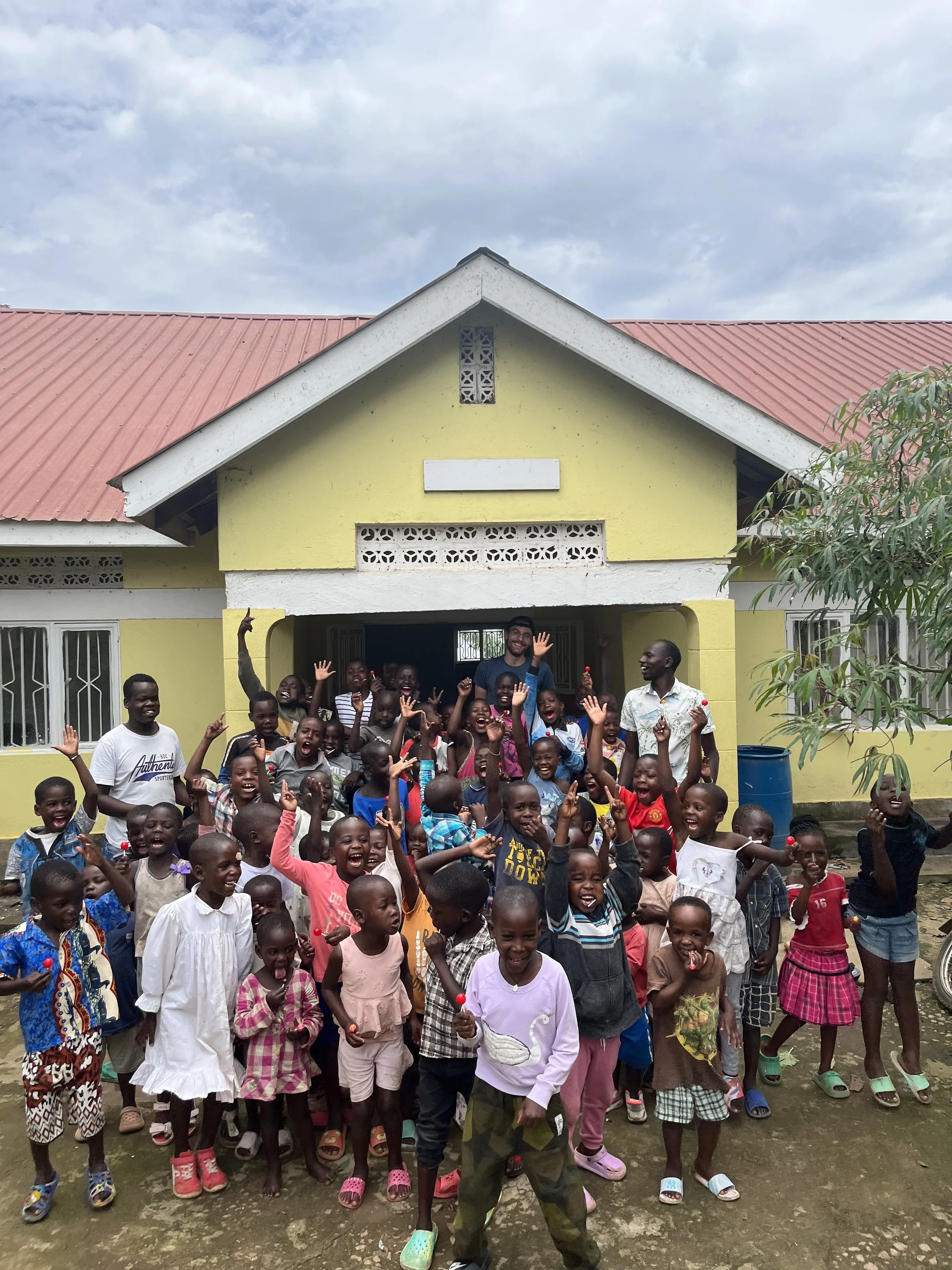 A group of children and adults standing outside a yellow house with a red roof, smiling and waving at the camera.