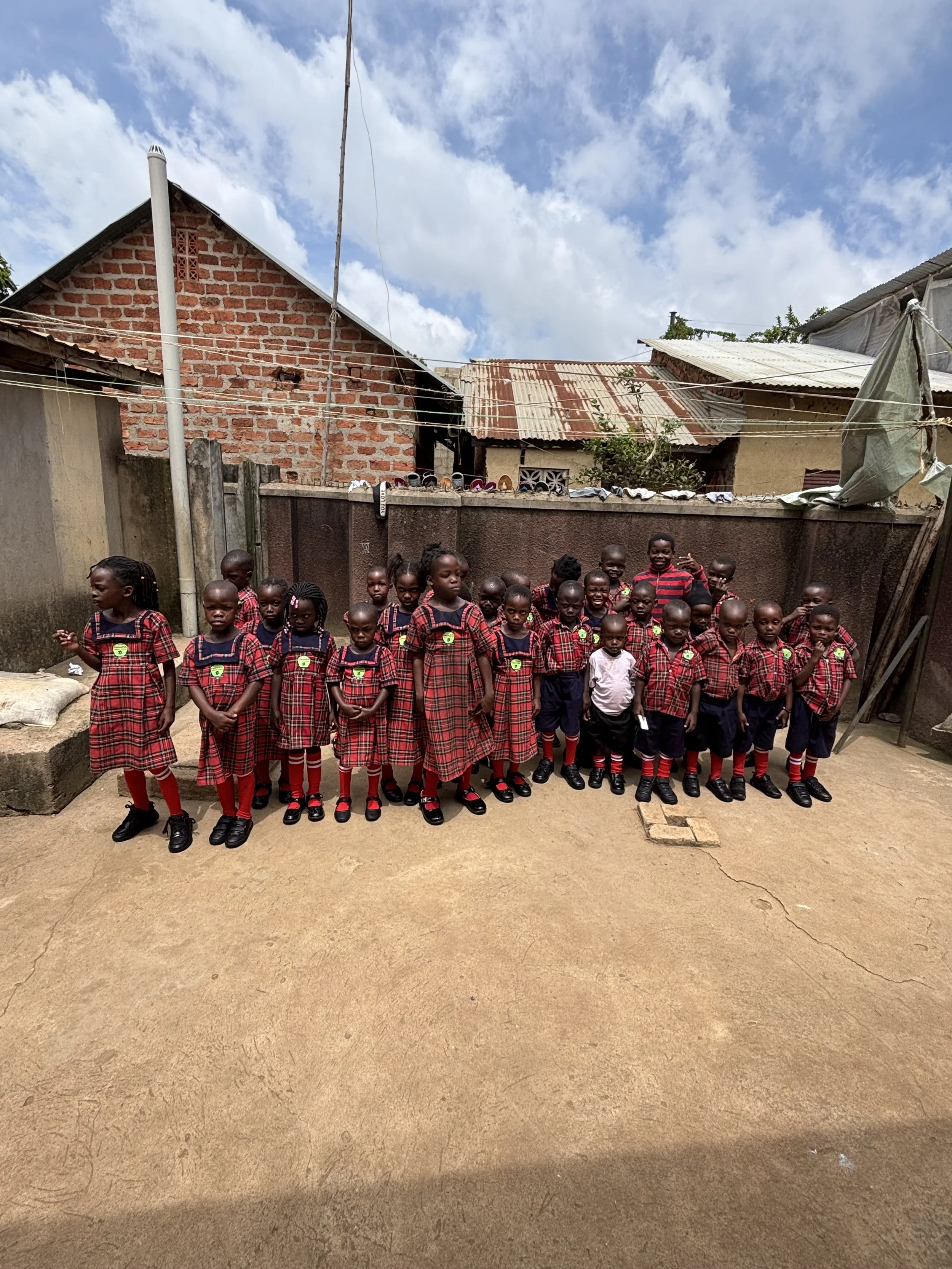 Group of young children in school uniforms standing outdoors in a courtyard with buildings and a cloudy sky in the background.