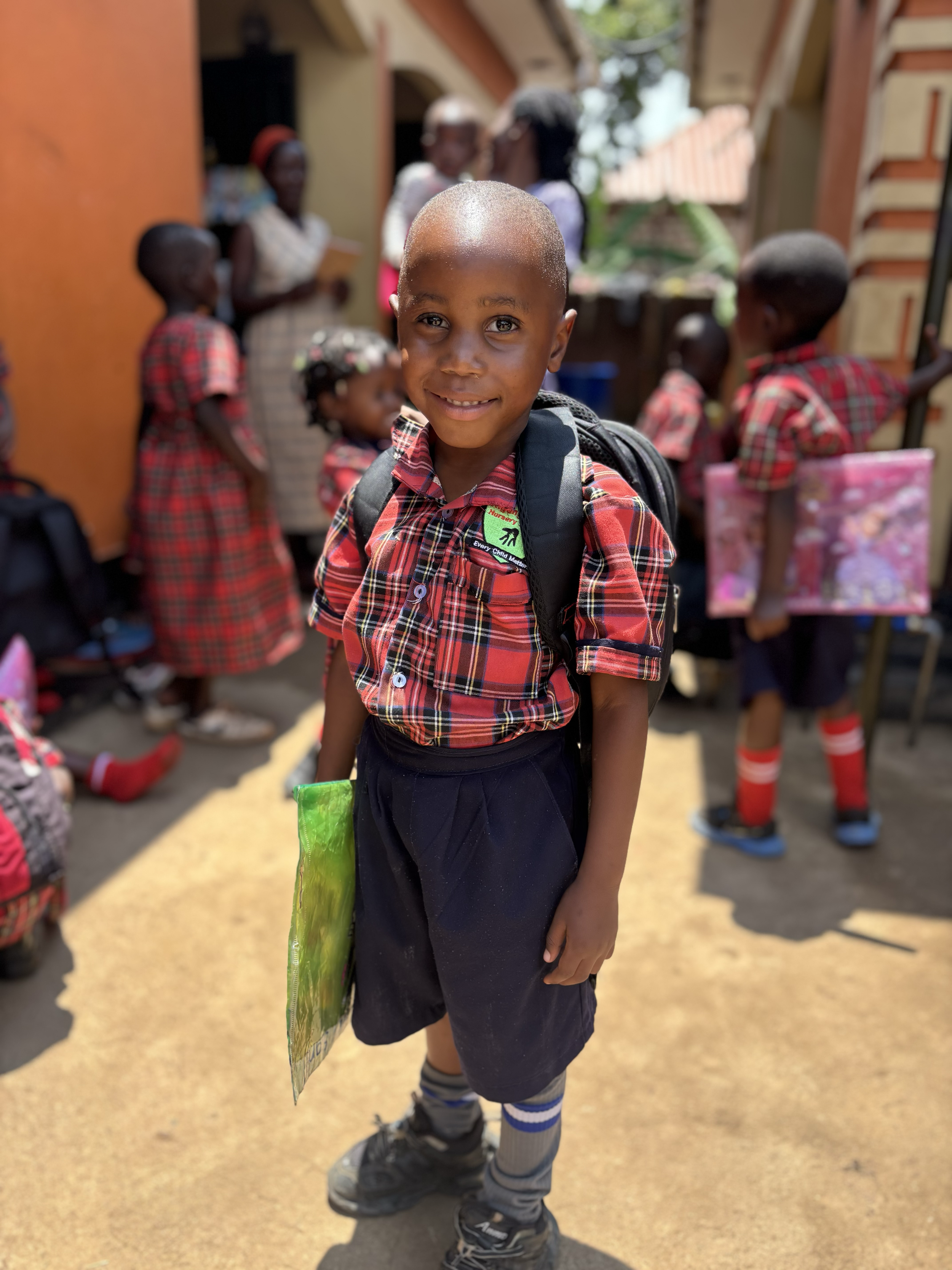 A young boy in a red plaid school uniform with a backpack smiling at the camera, with other children and adults in similar uniforms in the background at a school or community setting.