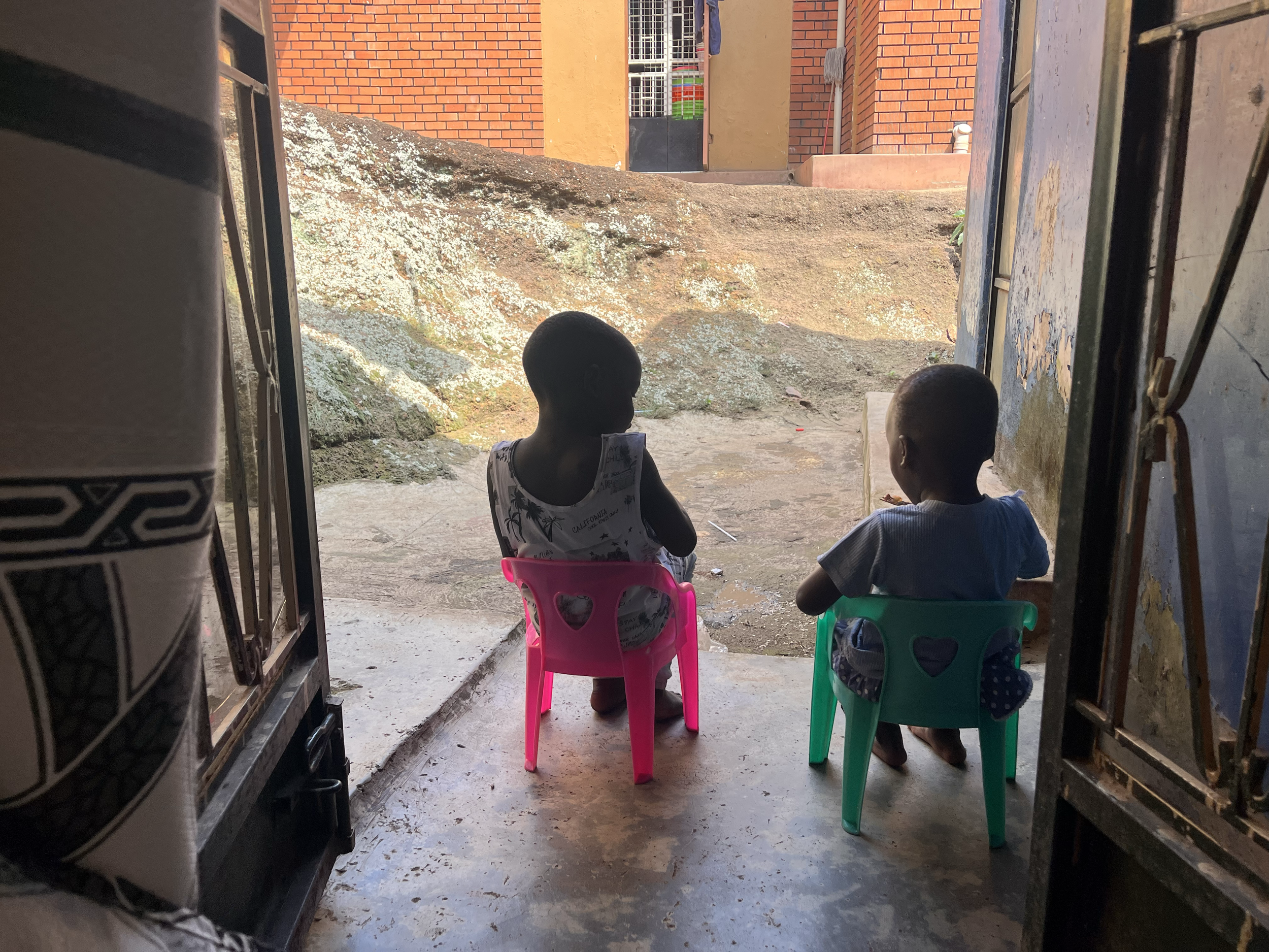 Two young children sit on small plastic chairs outside a doorway, facing away and looking towards an uneven, rocky outdoor area with a brown brick building in the background.