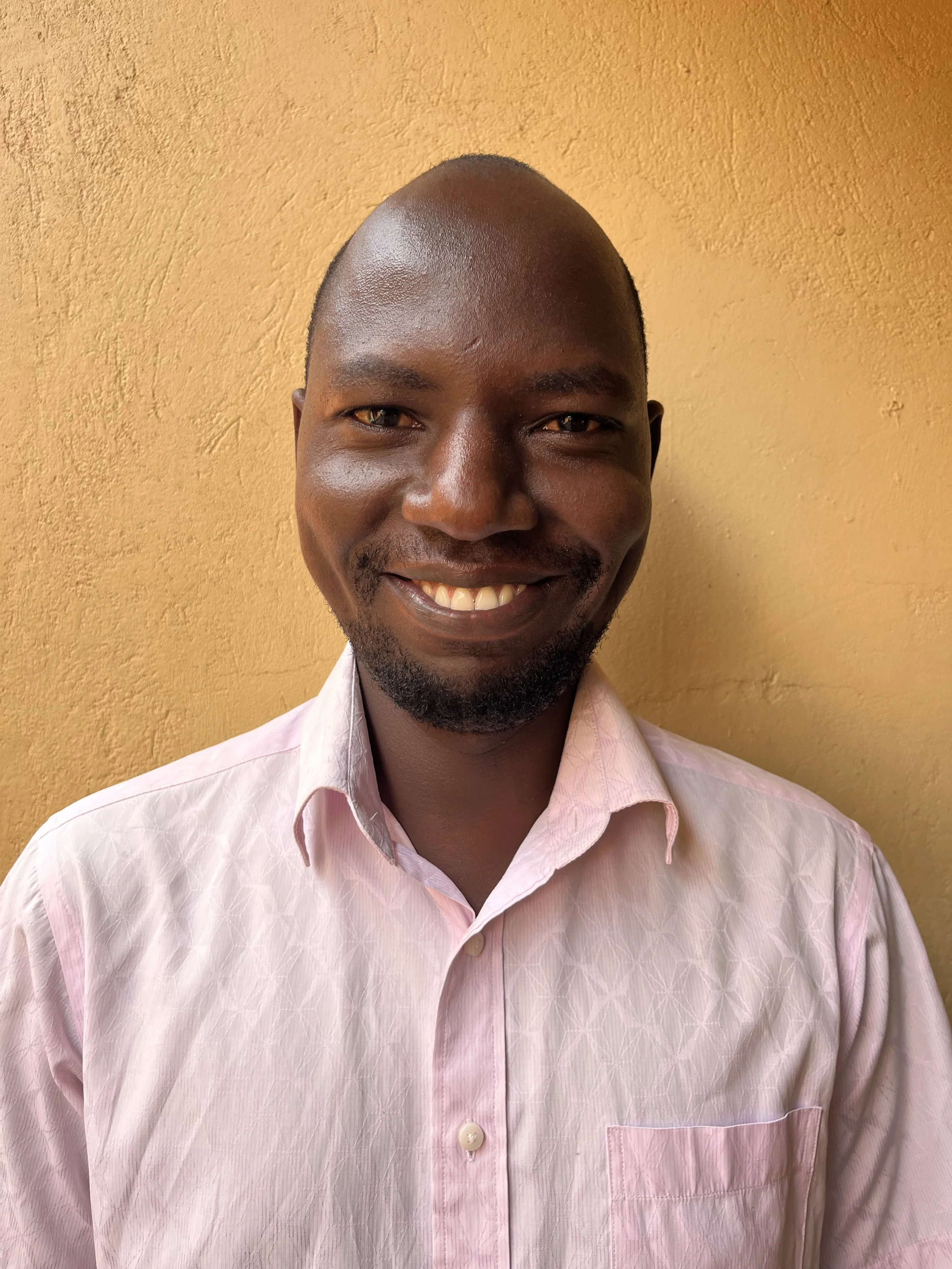 A smiling man with a shaved head and a beard is wearing a light pink button-up shirt and standing against a yellow textured wall.