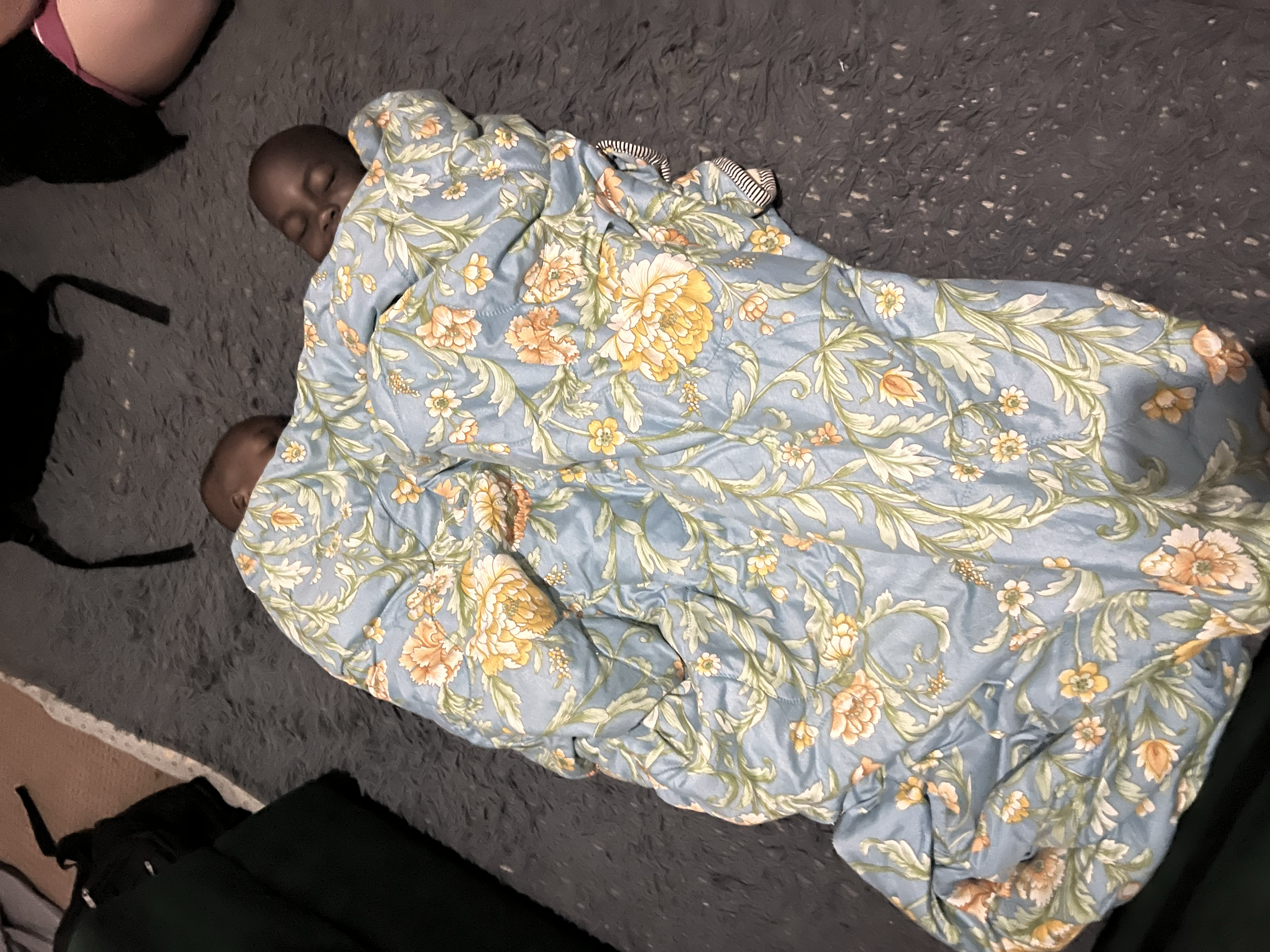 Three children sleeping on a dark gray carpeted floor, covered with a light blue floral blanket, with two of their faces visible and one face partially covered.