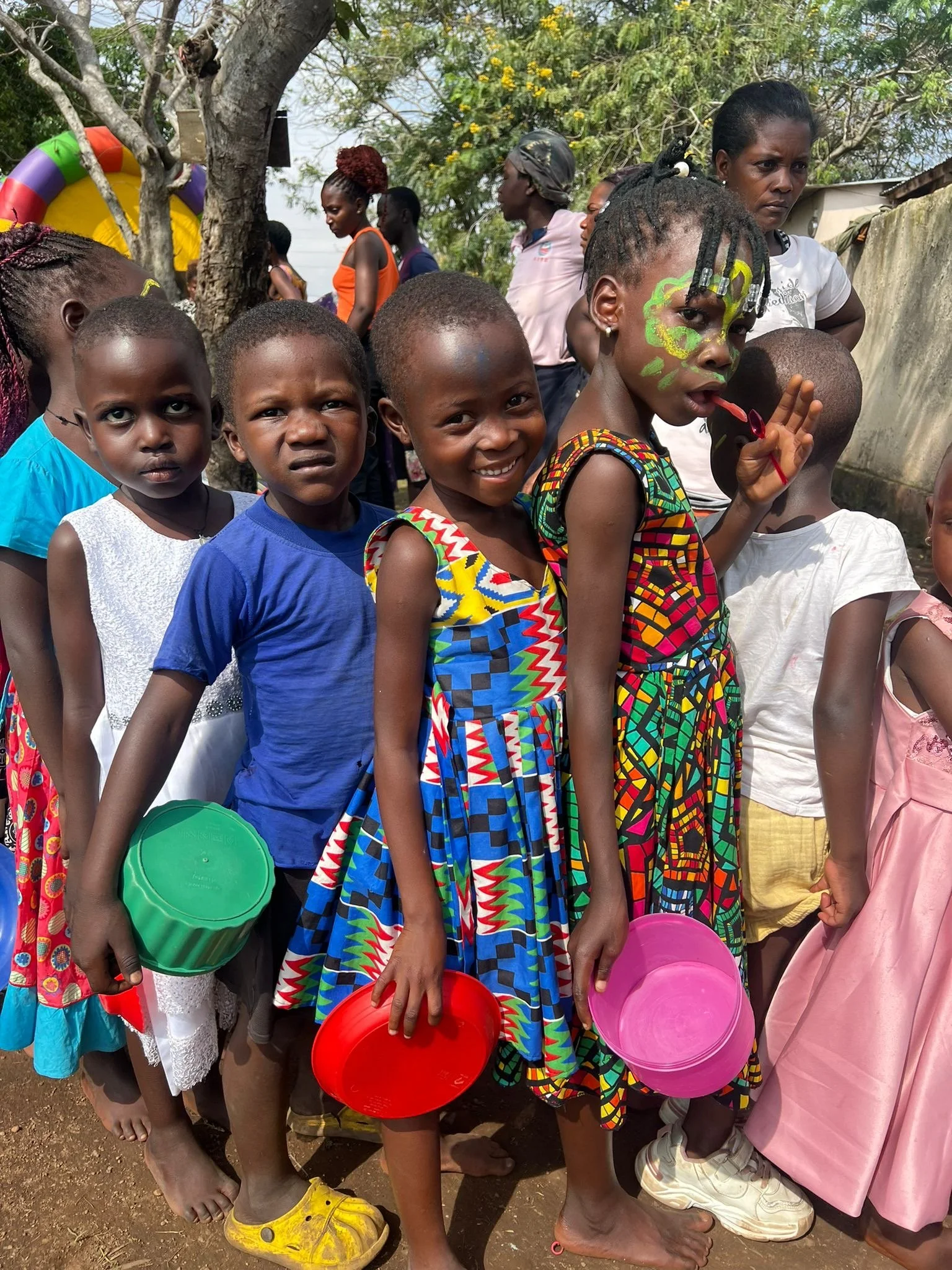 Group of young children at an outdoor event, some smiling and holding colorful bowls, with adults and trees in the background.
