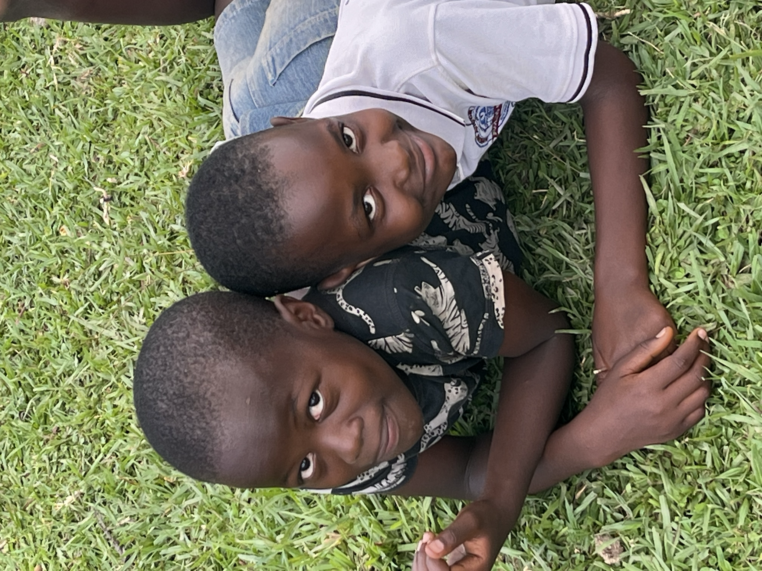 Two young boys lying on grass, facing the camera, looking up with relaxed expressions.