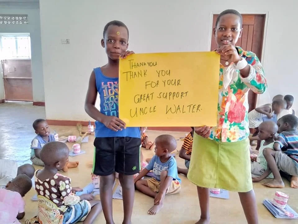 Two children standing and holding a yellow thank you sign, with several children sitting on the floor in the background.