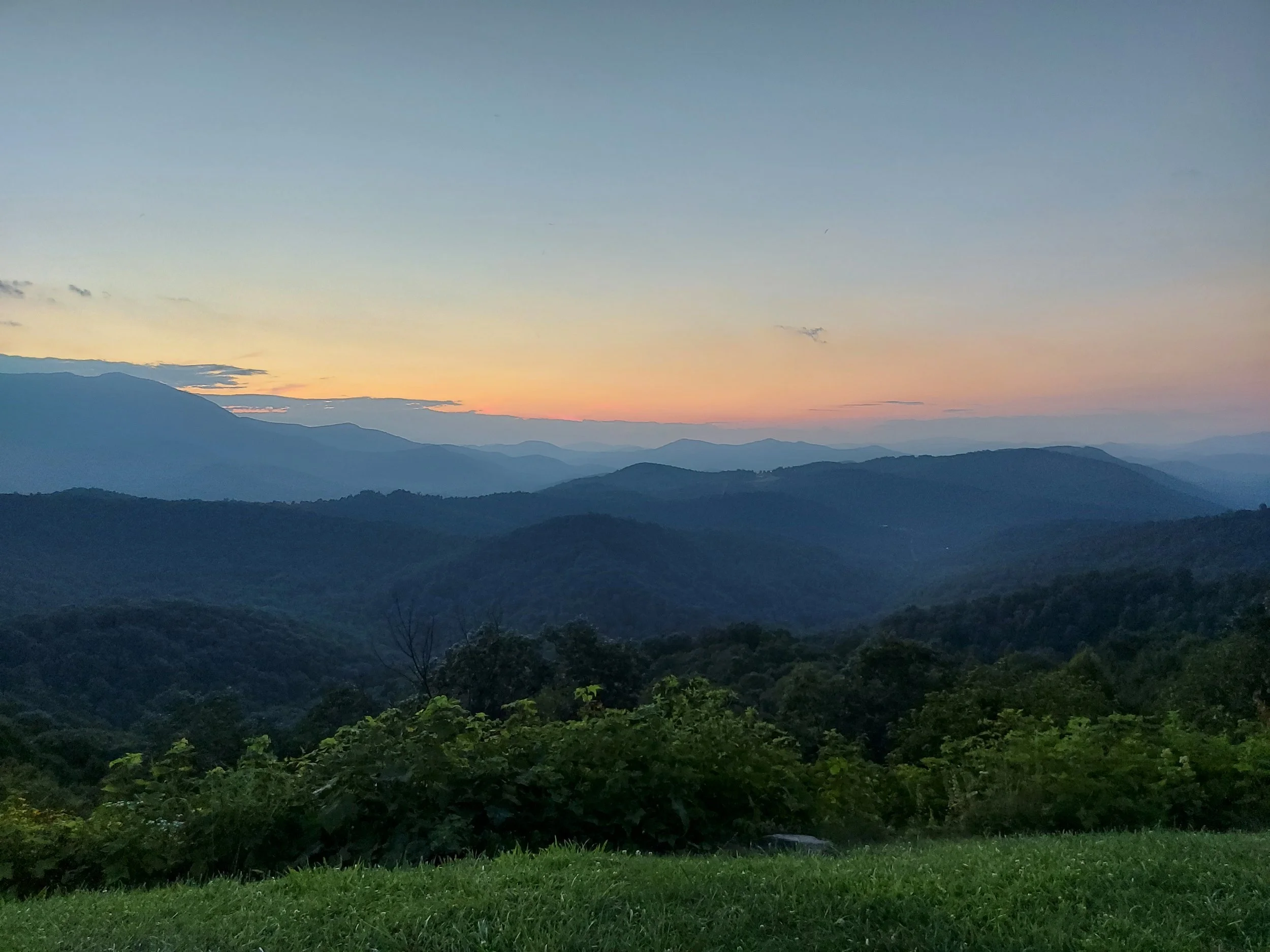 Scenic view of layered mountains at sunset, with a clear sky and lush green foreground.