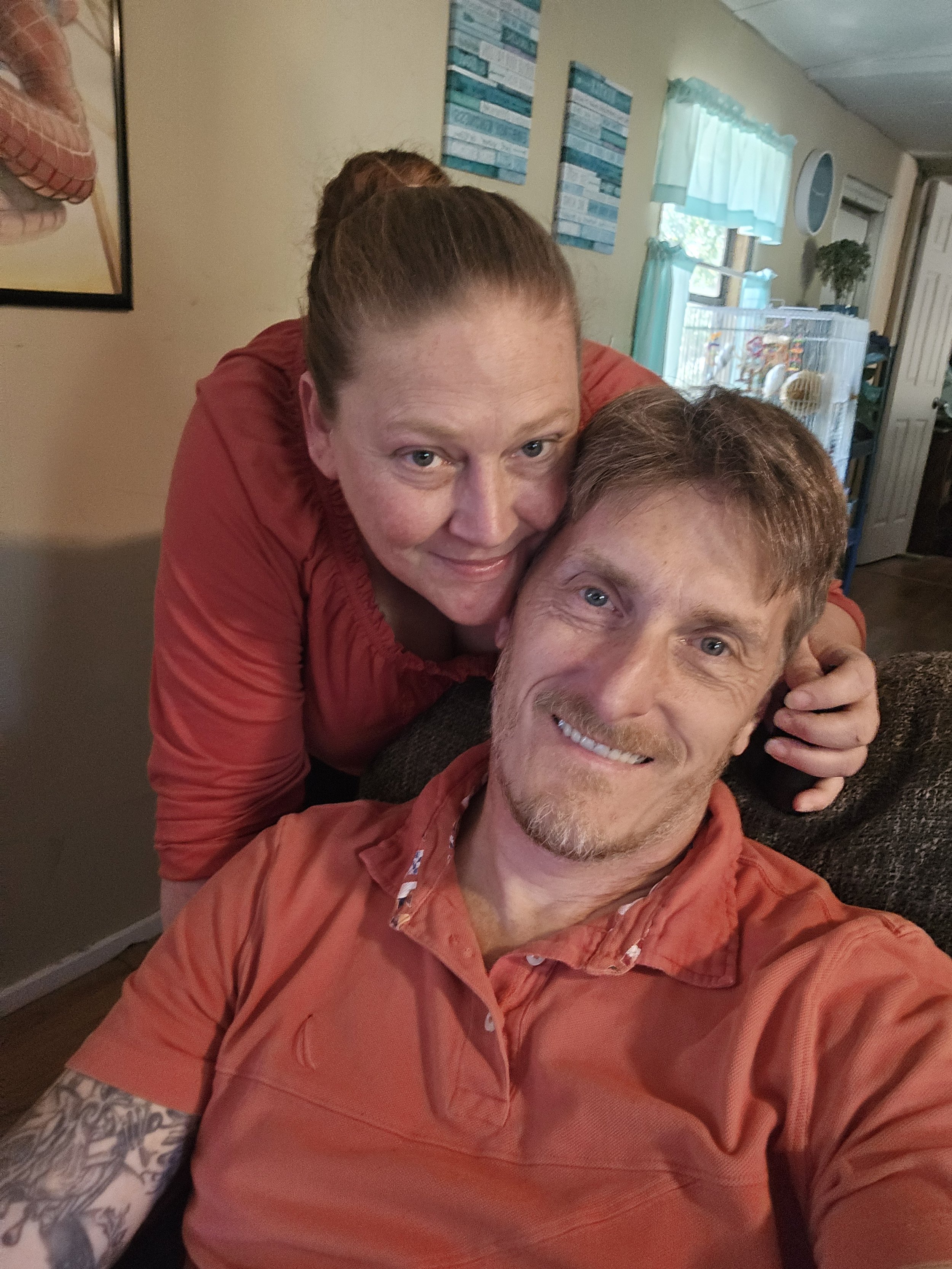 A smiling couple taking a selfie inside a house. The woman is leaning over the man, both wearing red shirts. The background shows a window with a curtain and some artwork on the wall.