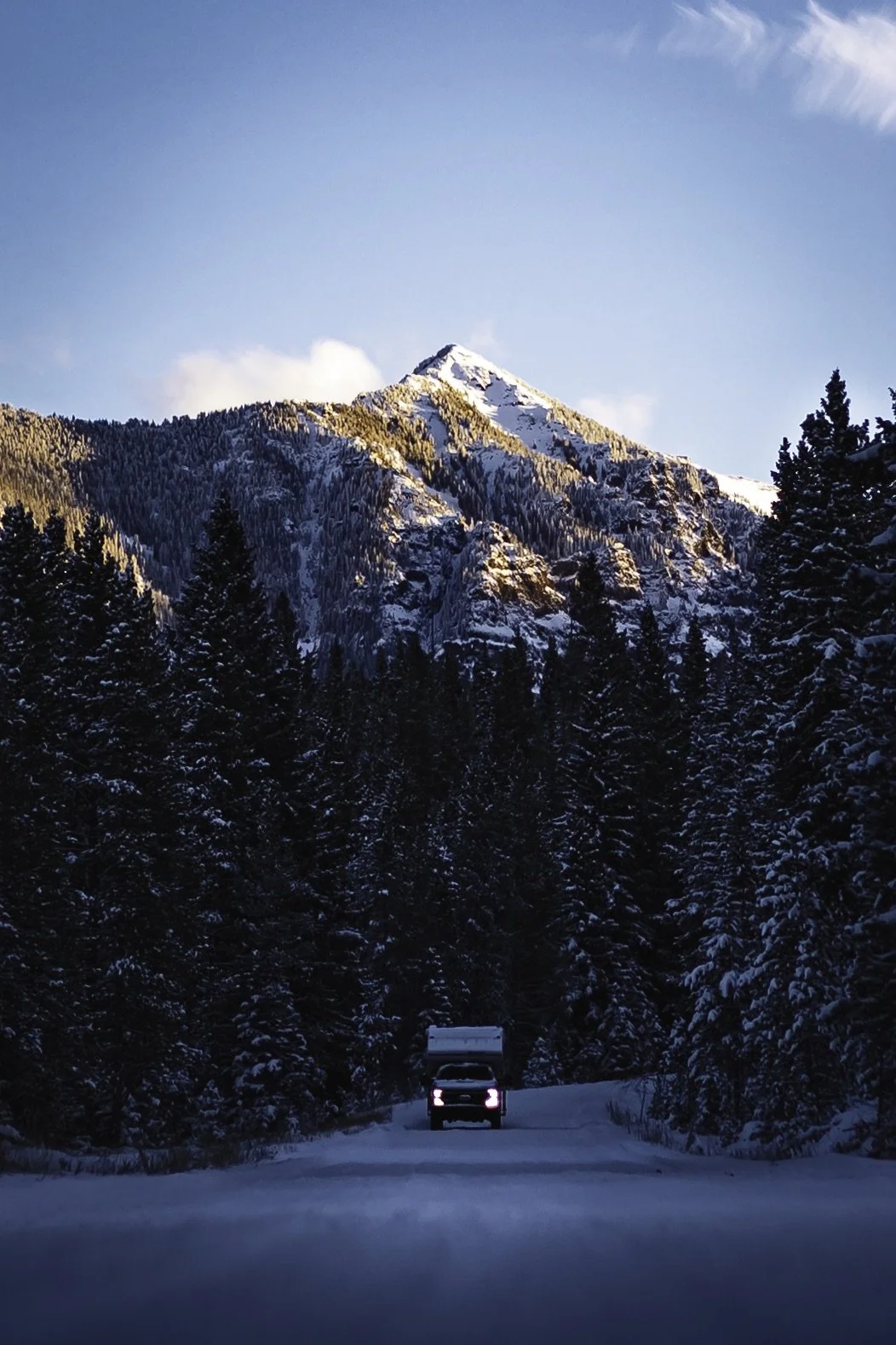 A snowy mountain landscape with a forest of snow-covered trees in the foreground, a vehicle on a snow-covered road, and the mountain peak illuminated by sunlight under a blue sky.
