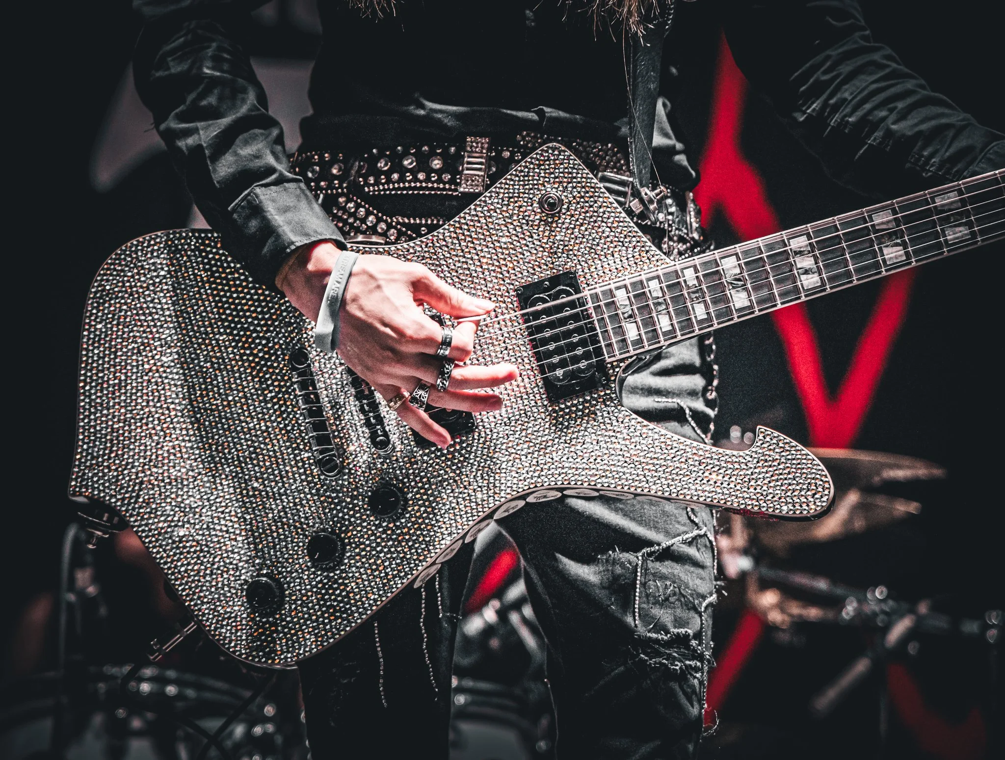 Close-up of a musician playing an electric guitar covered in rhinestones, wearing a black leather jacket and ripped black jeans, with a red background.